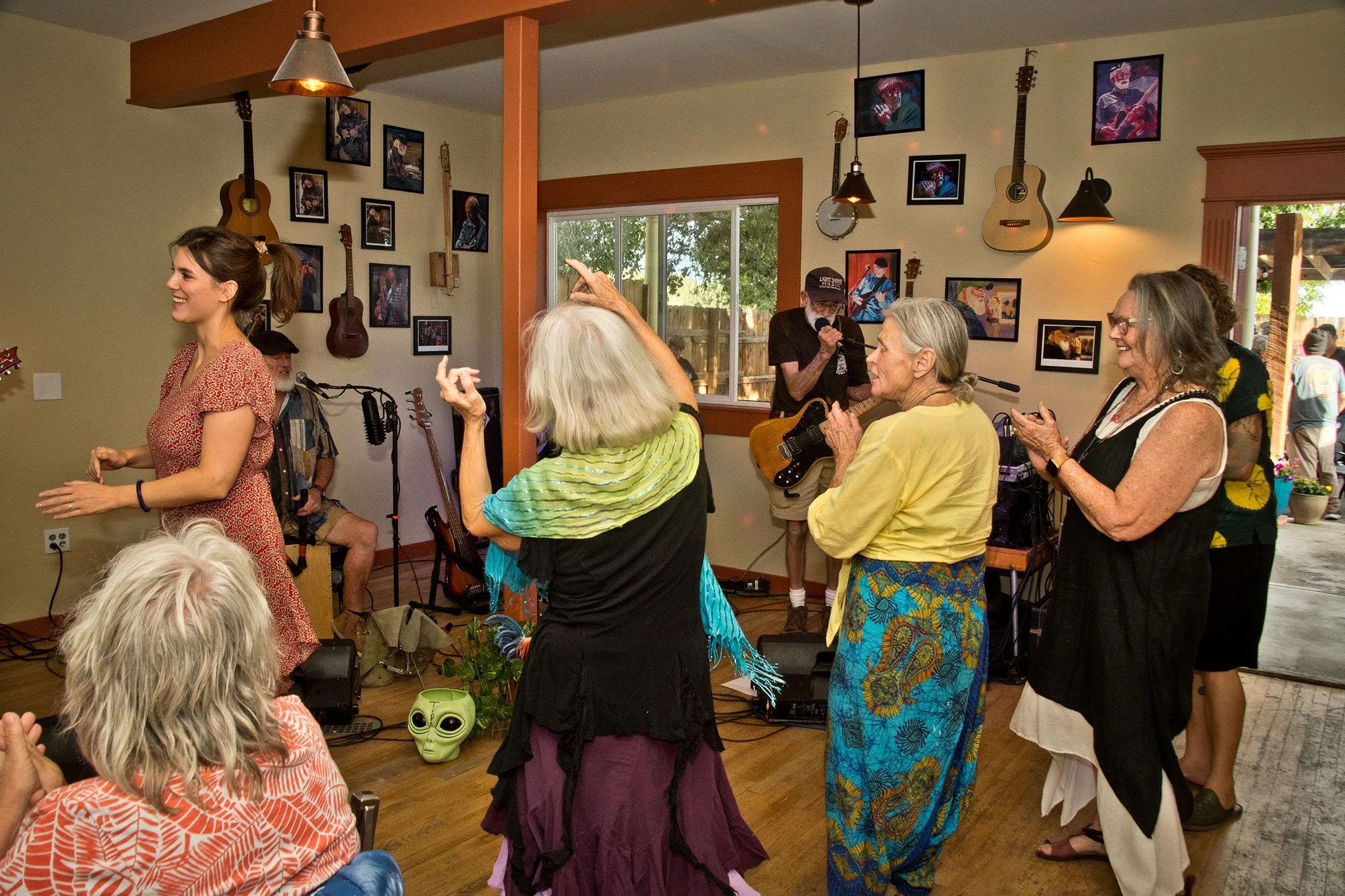 A group of people at an indoor music gathering, with some dancing and others playing instruments, in a room decorated with framed photos and guitars on the wall.
