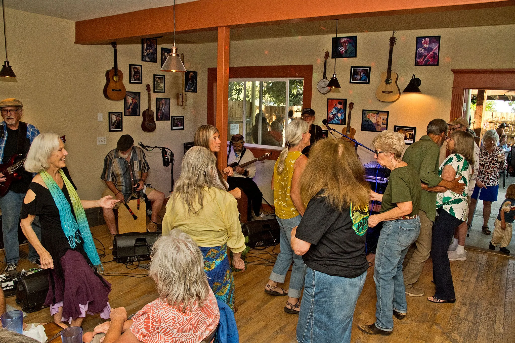 People dancing and socializing in a cozy indoor space with guitars and framed photographs on the walls, during a live music event.