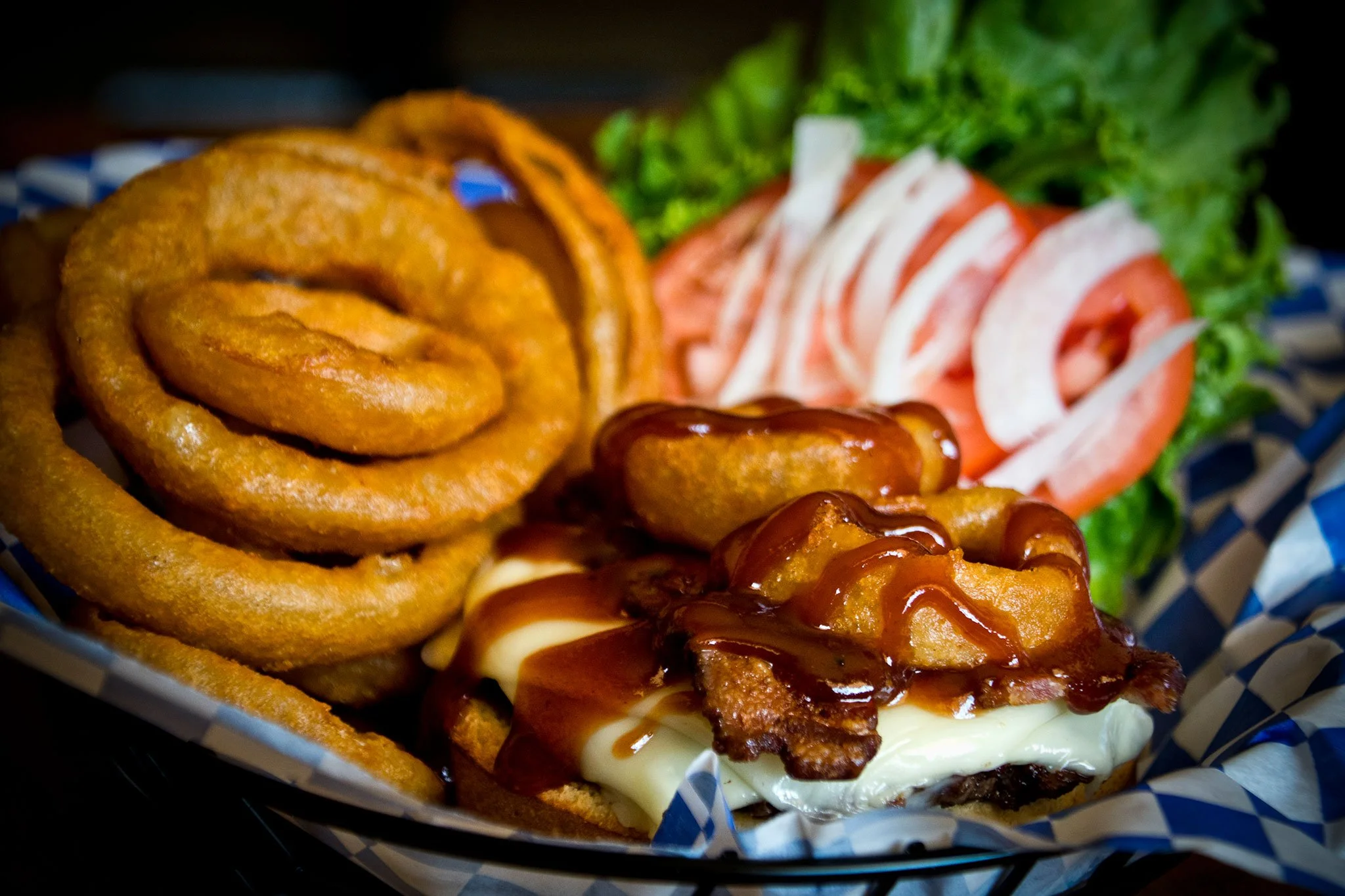 A close-up of a burger with onion rings and barbecue sauce, garnished with lettuce and tomato slices, served in a paper-lined basket.