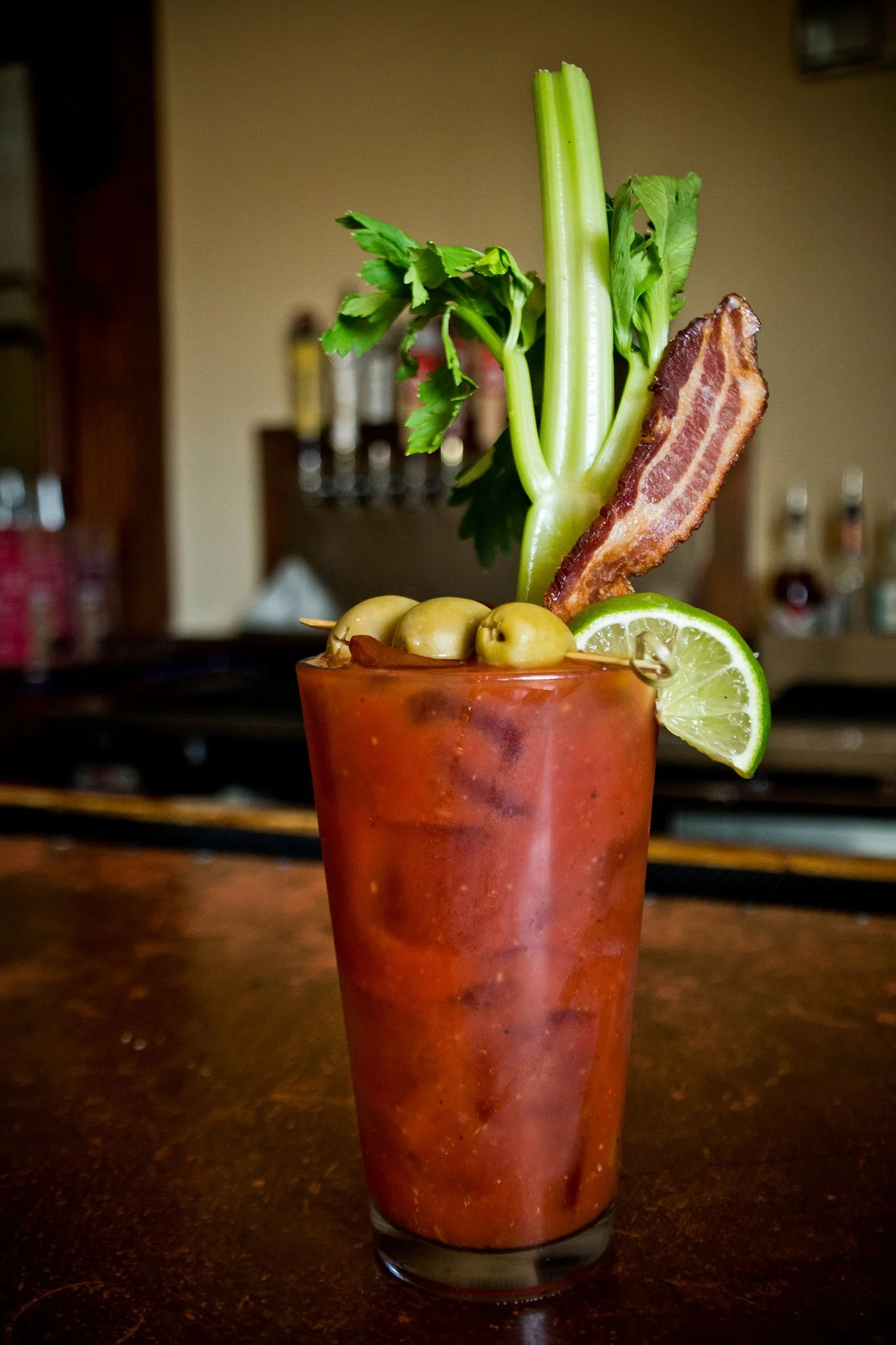 A Bloody Mary cocktail garnished with celery stalk, bacon strip, lime wedge, green olives, and parsley in a tall glass.