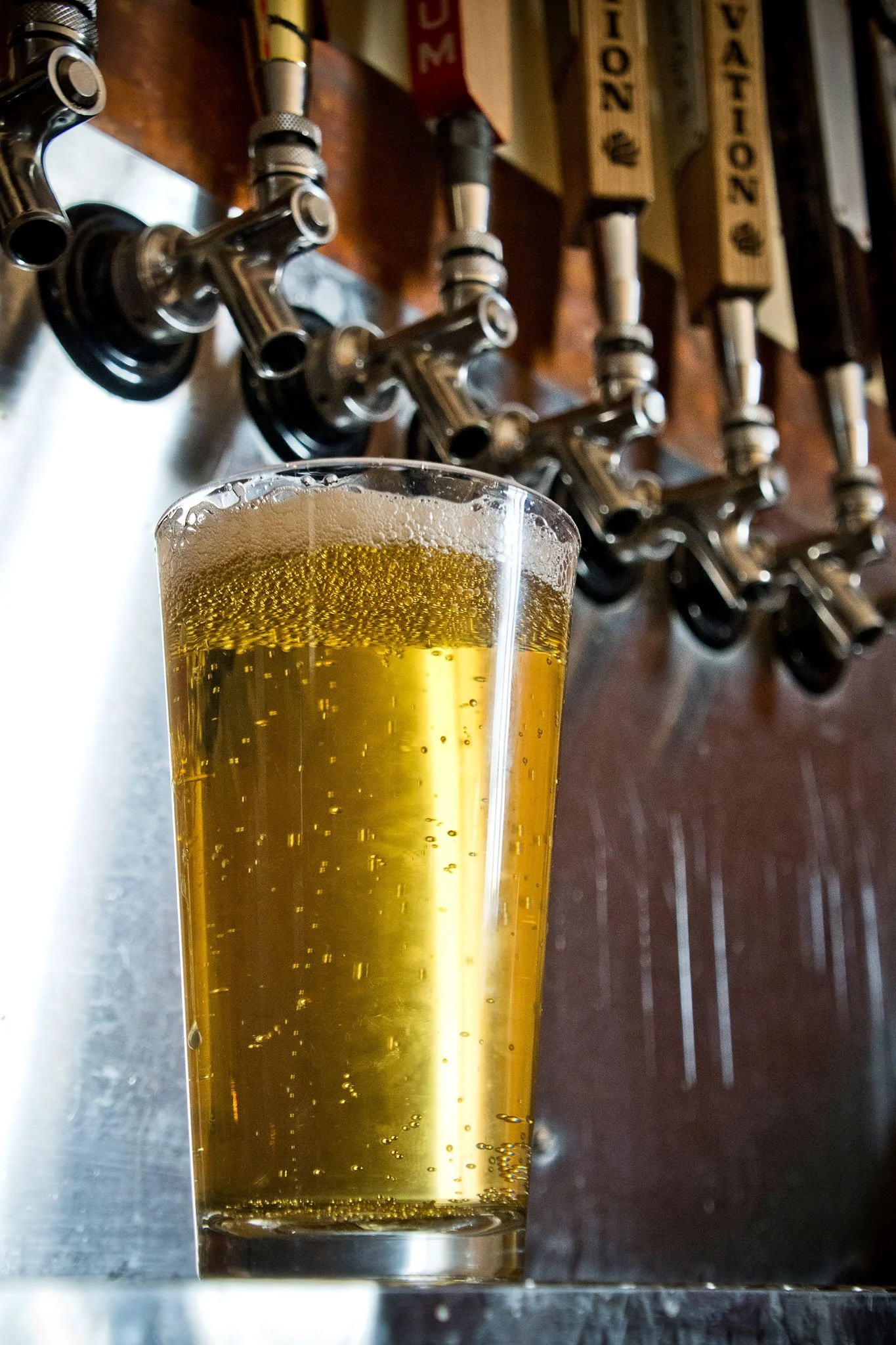 A glass of beer filled with golden liquid and bubbles, placed on a metallic surface with beer taps and wooden signs in the background.