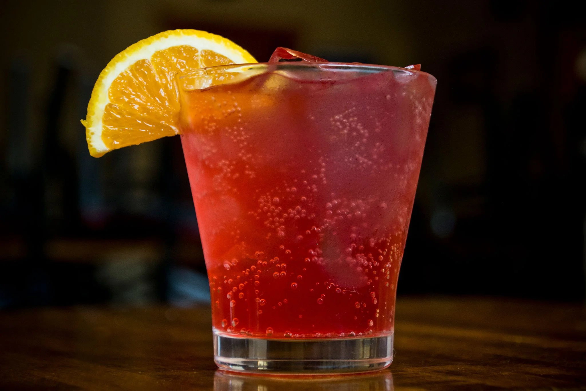 Red soda in a glass with bubbles, garnished with an orange slice on the rim.
