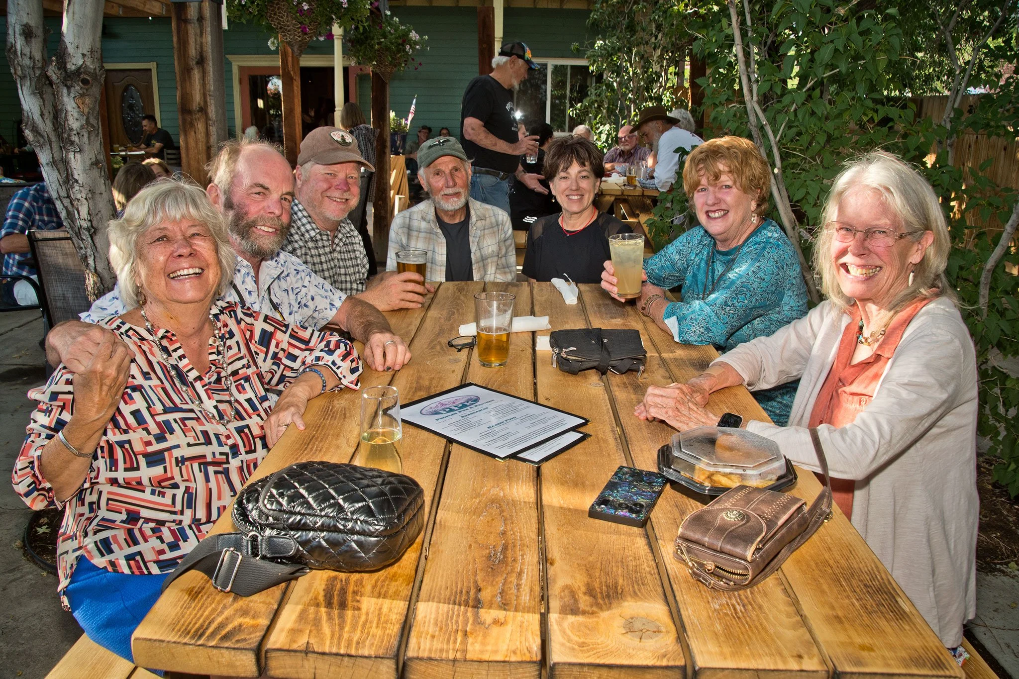 A group of seven older adults, four women and three men, enjoying drinks and smiling at an outdoor restaurant or patio. They are seated around a wooden table with menus, purses, and drinks. The background shows other diners, trees, and a green buildi