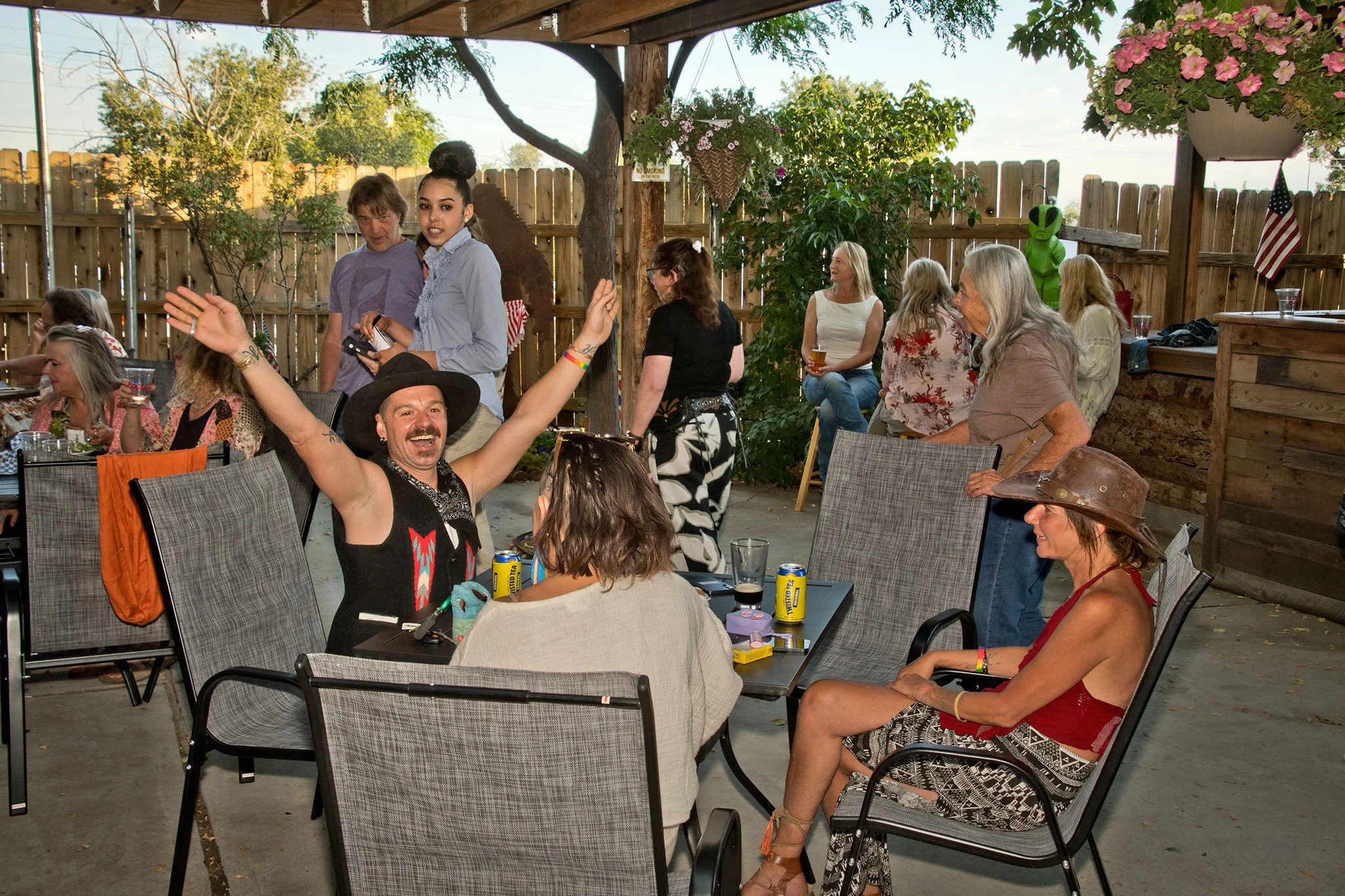 People gathered in an outdoor patio enjoying a social event with some holding drinks, seated and standing around tables, with a wooden fence, trees, and decorative plants in the background.
