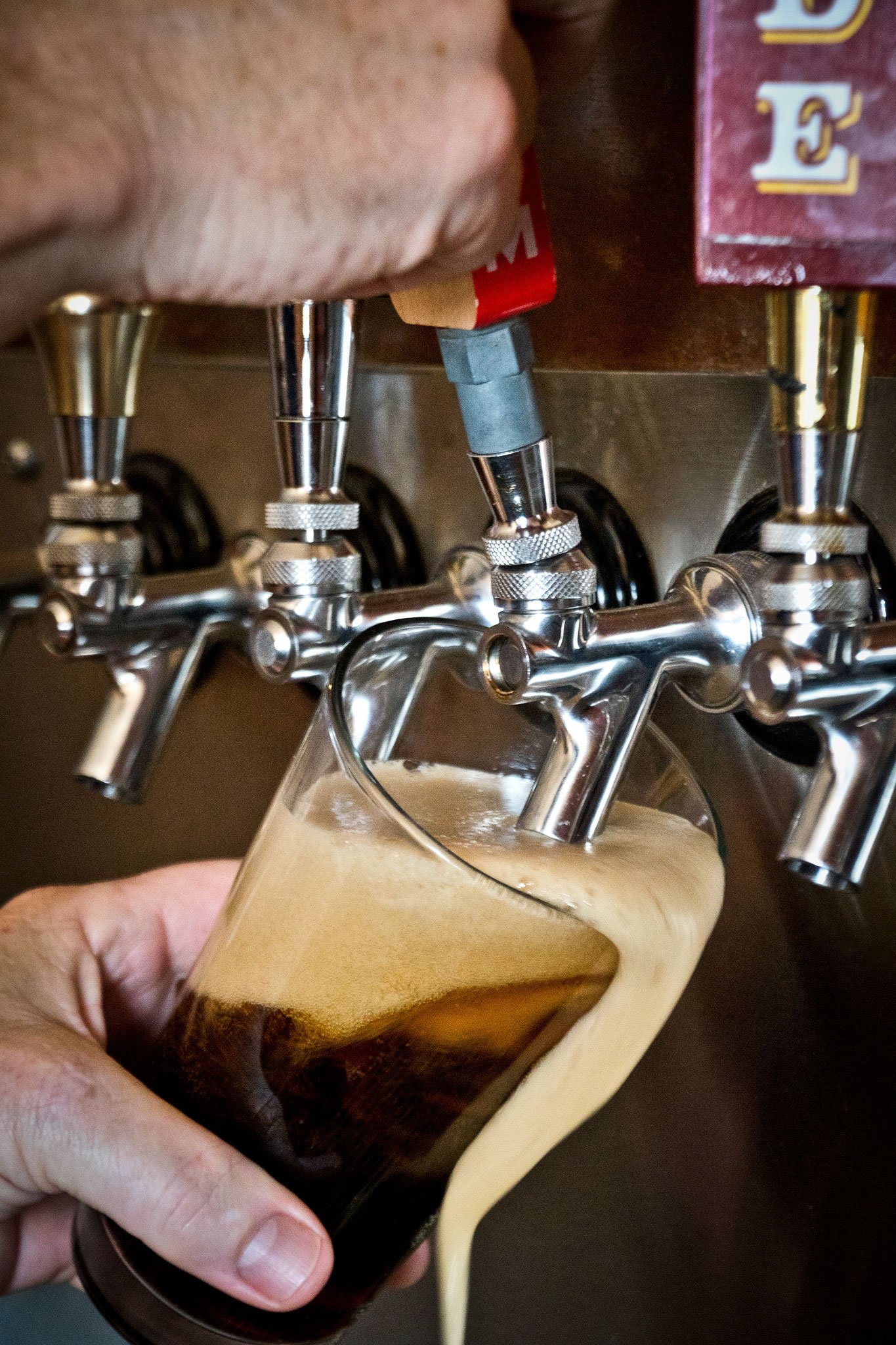 A person is filling a glass with a dark beverage from a beer tap that has multiple spouts, with a wooden wall and a red box nearby.