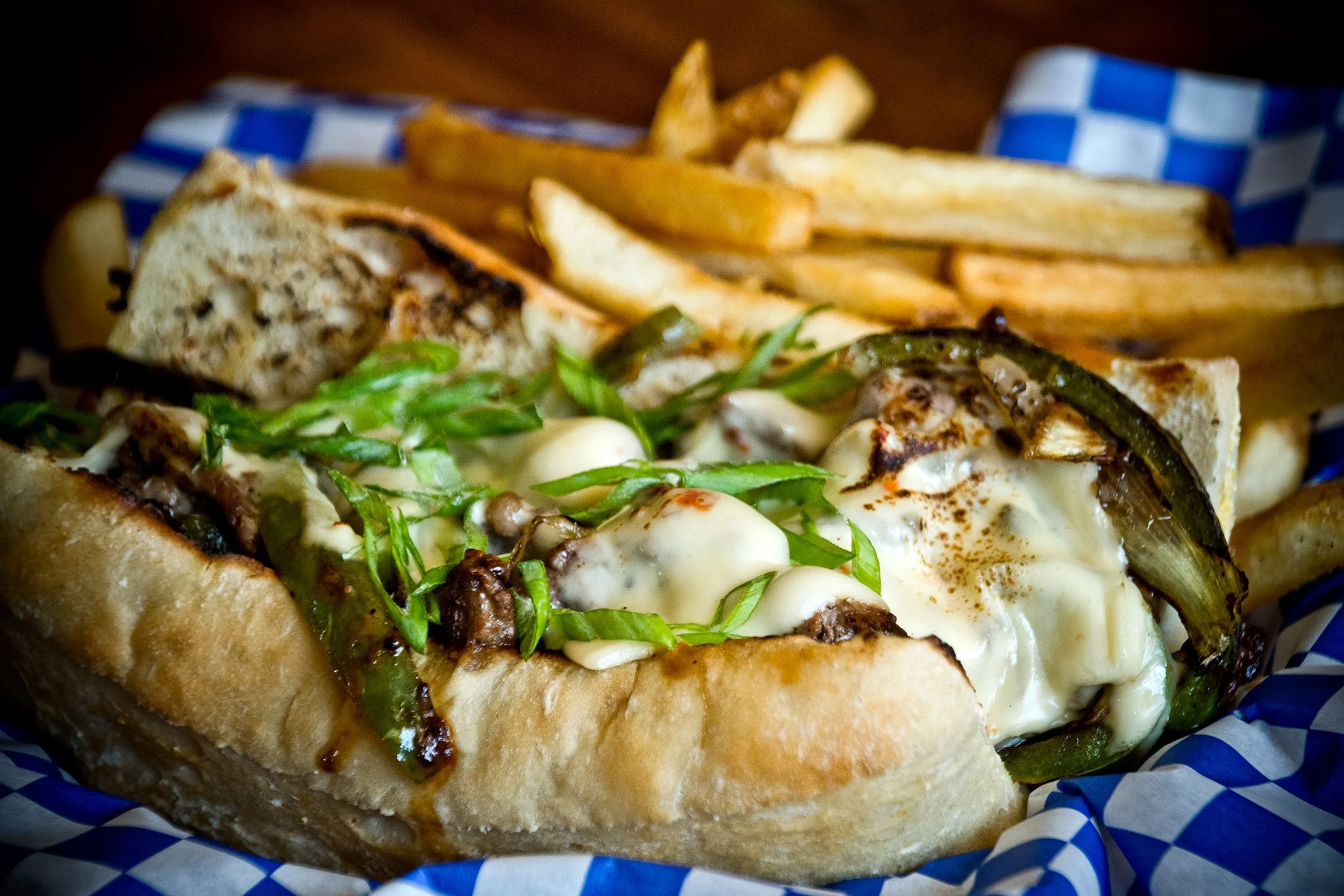 Close-up of a sandwich with grilled vegetables, melted cheese, and fresh greens in a sandwich bun, served with French fries on a checkered paper tray.