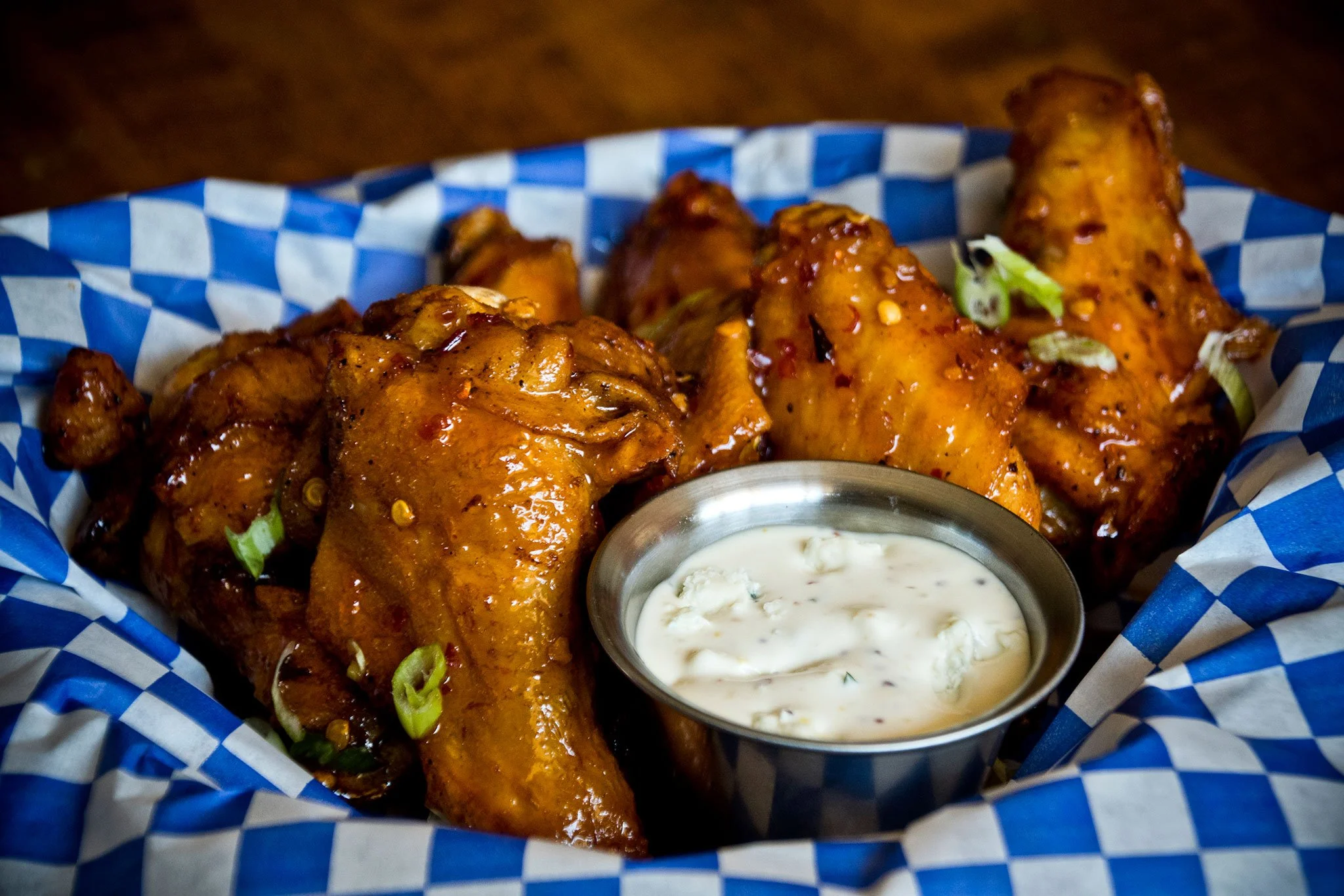 Close-up of buffalo chicken wings served in a basket with blue checkered paper, accompanied by a small container of ranch dressing