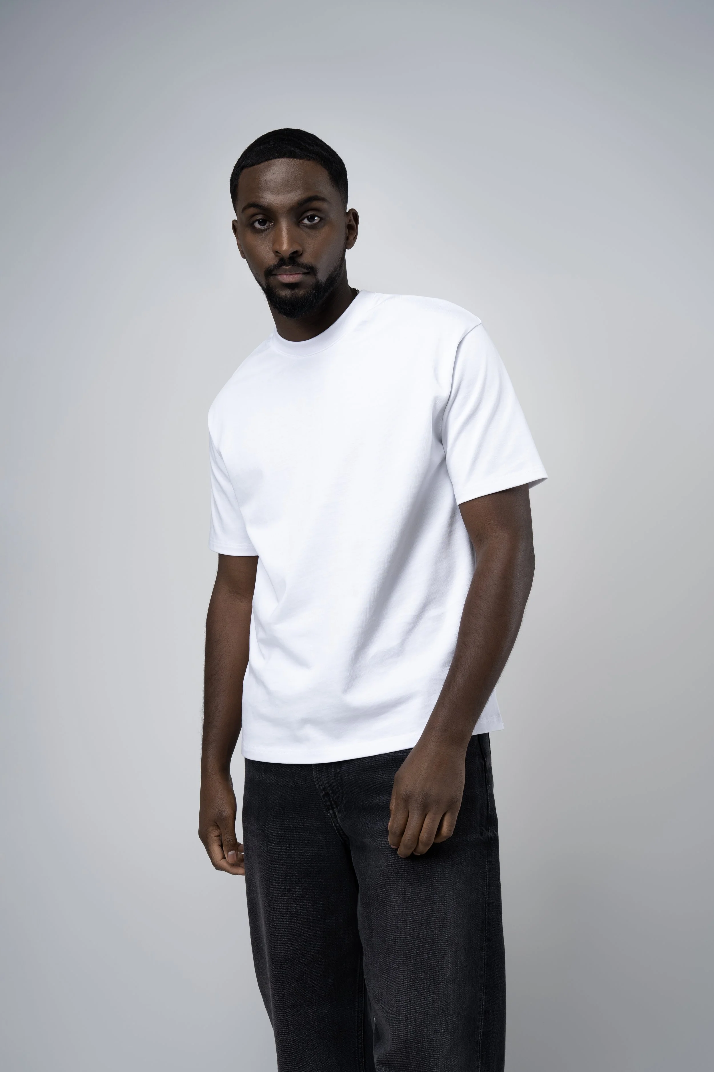 Young man with dark skin, short black hair, and a beard, wearing a white t-shirt and black jeans, standing against a plain light gray background.