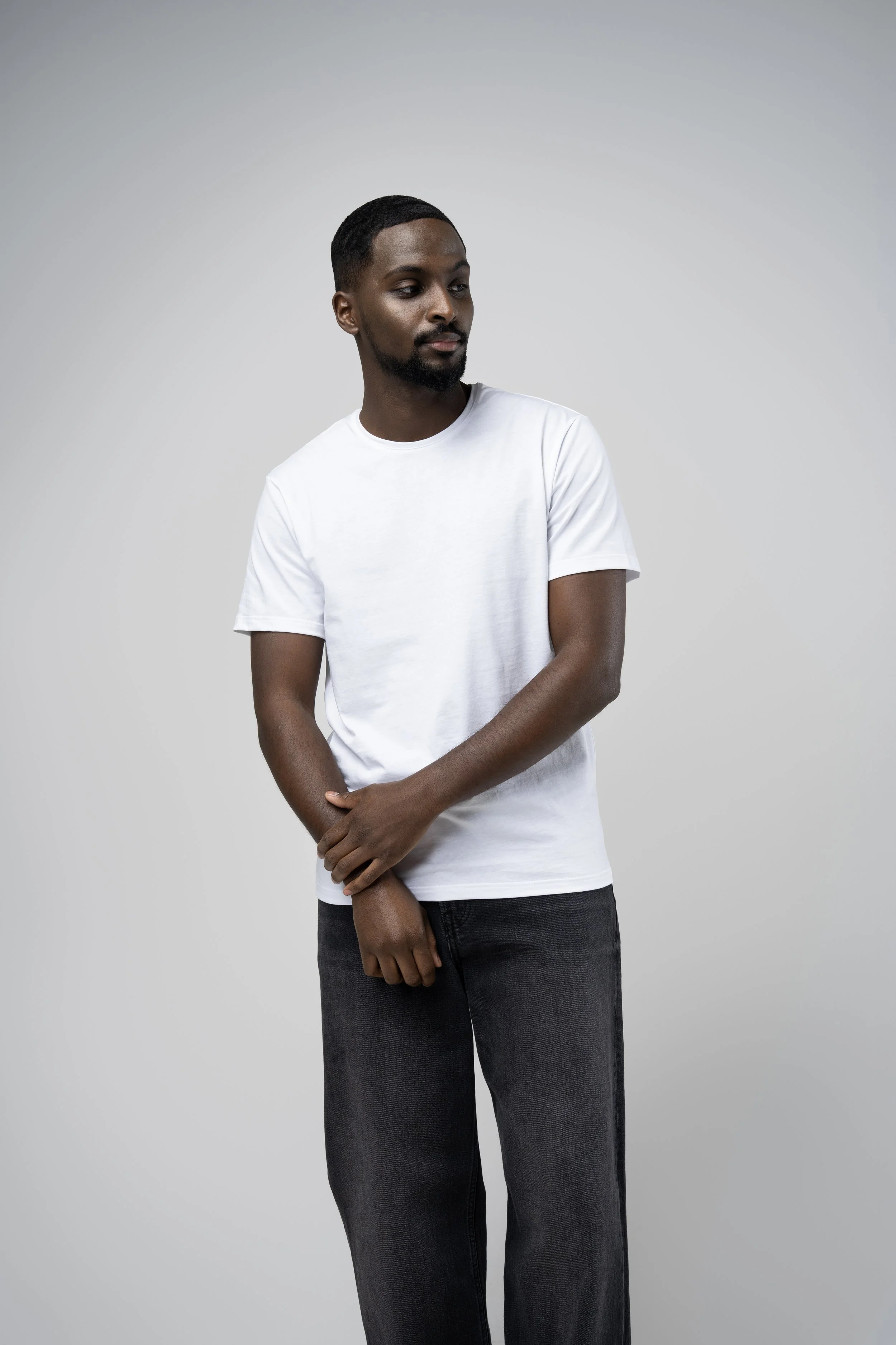 A young man with a beard and short hair standing against a plain gray background, wearing a white t-shirt and black jeans.
