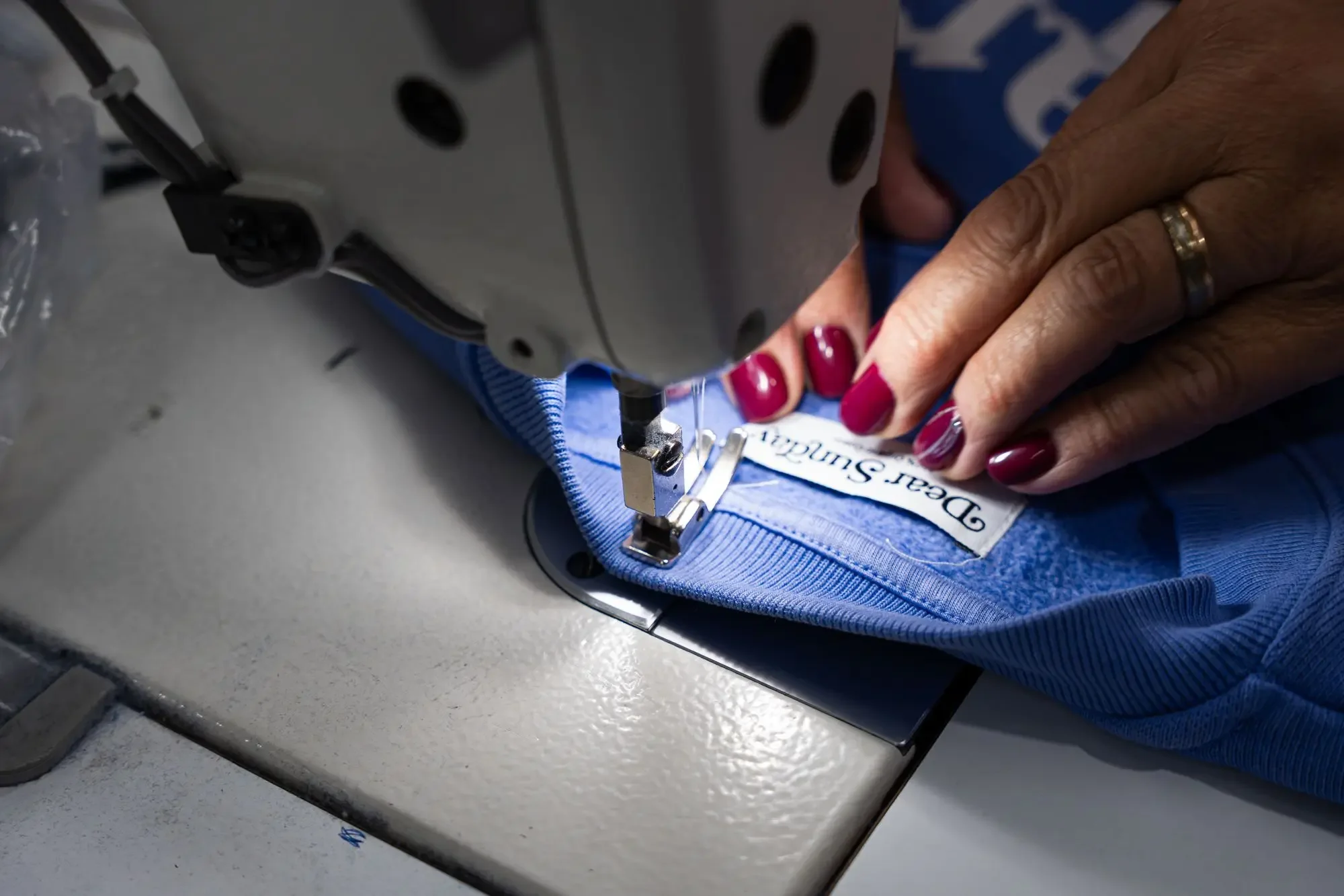 Close-up of a person sewing a blue fabric with a sewing machine, showing their hands with painted nails and a wedding ring, and a sewing label reading 'Dear Sunchild'.