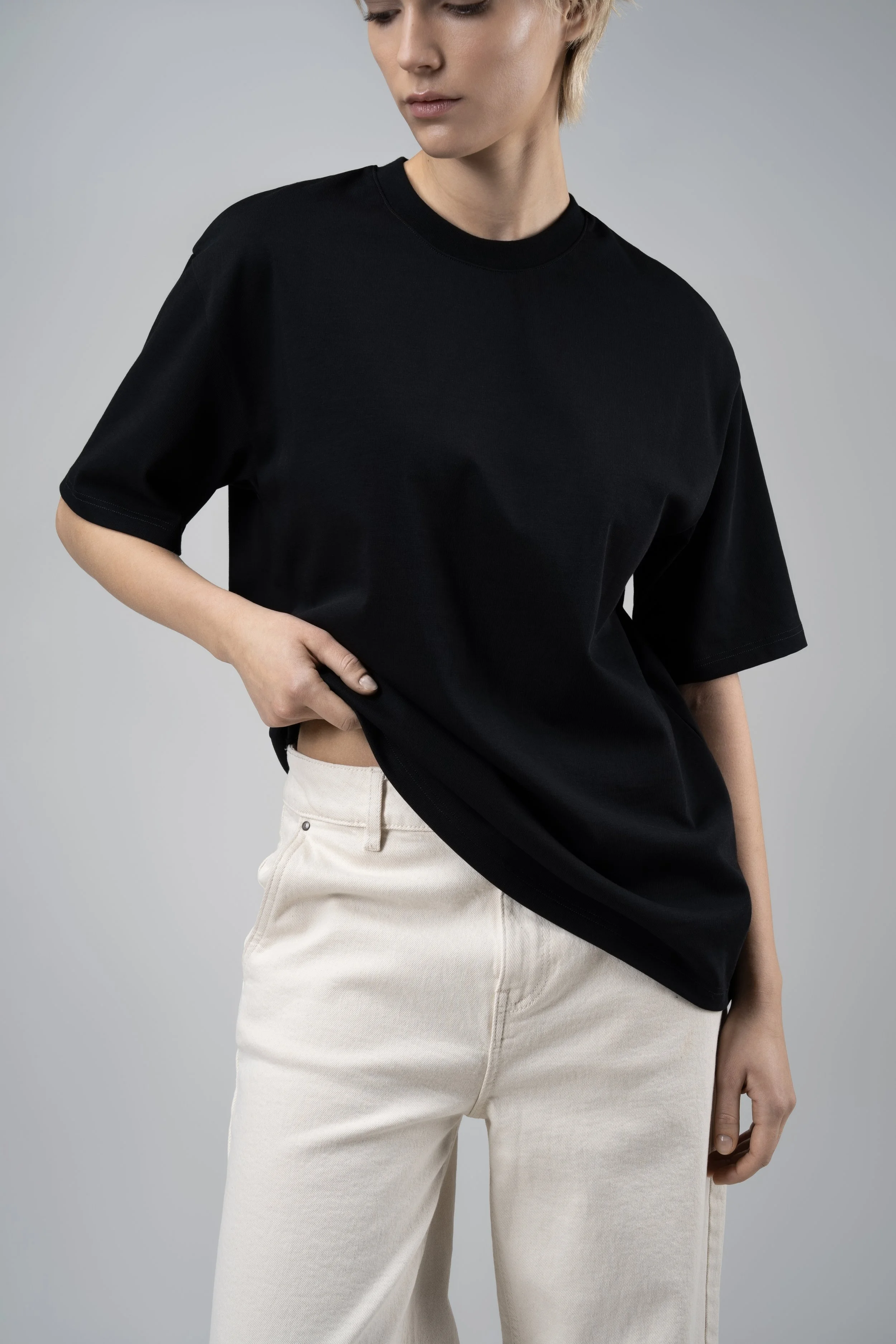 A woman wearing a black t-shirt and beige pants, with her left hand resting on her hip, against a neutral gray background.