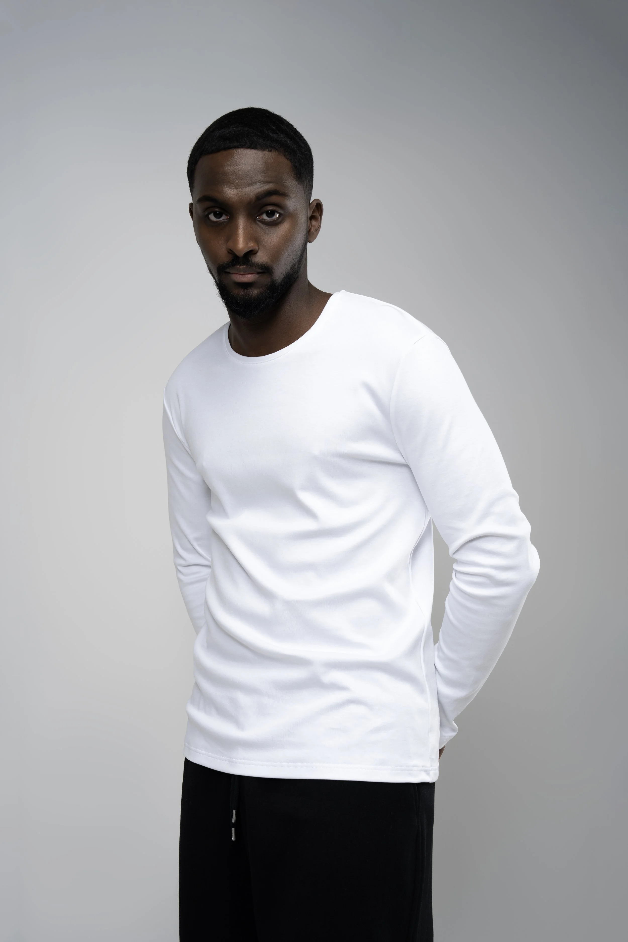 A man with dark skin, short hair, and a beard wearing a white long-sleeve shirt and black pants, standing with one hand behind his back against a plain gray background.