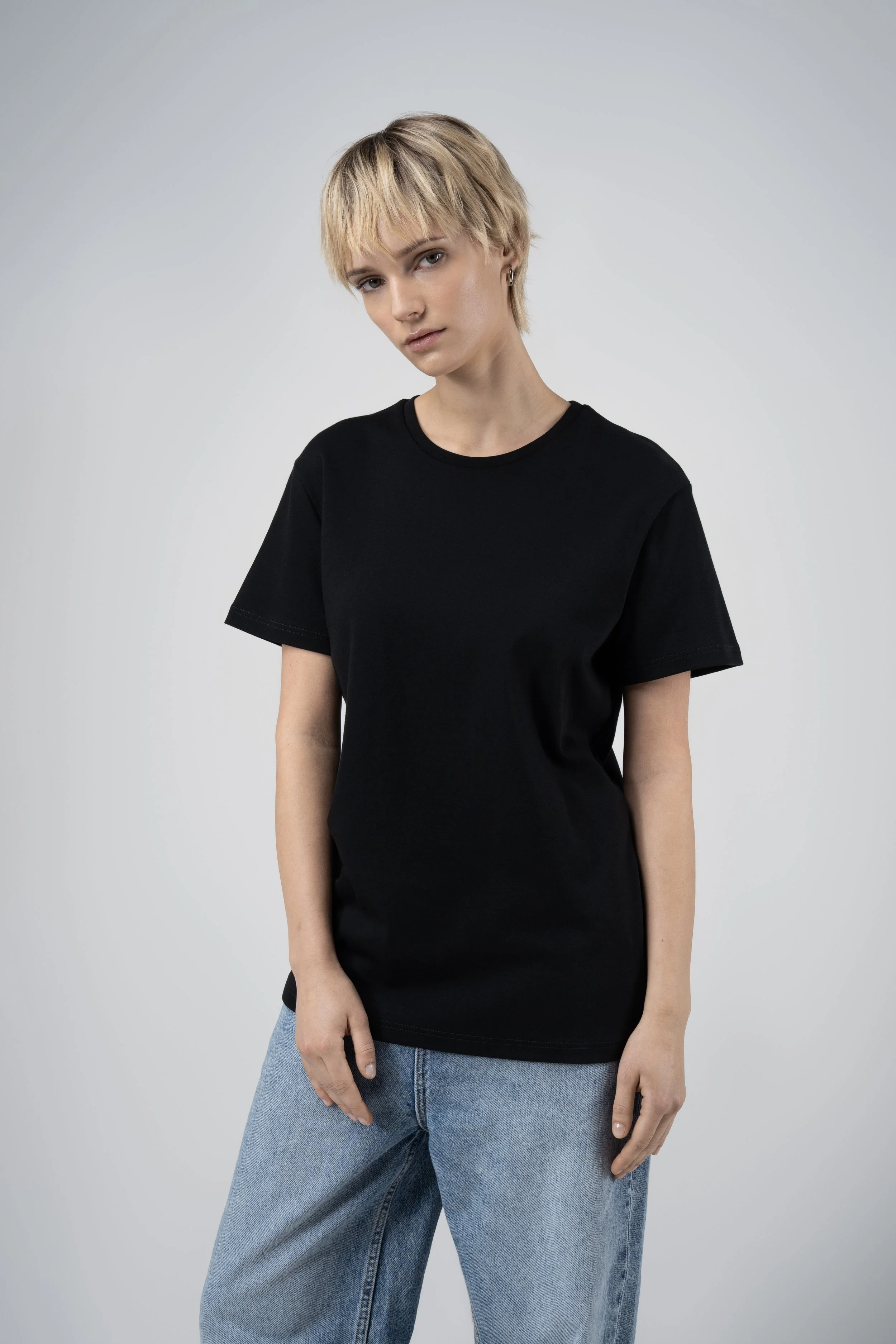 A woman with short blonde hair, wearing a black t-shirt and blue jeans, standing against a plain gray background.