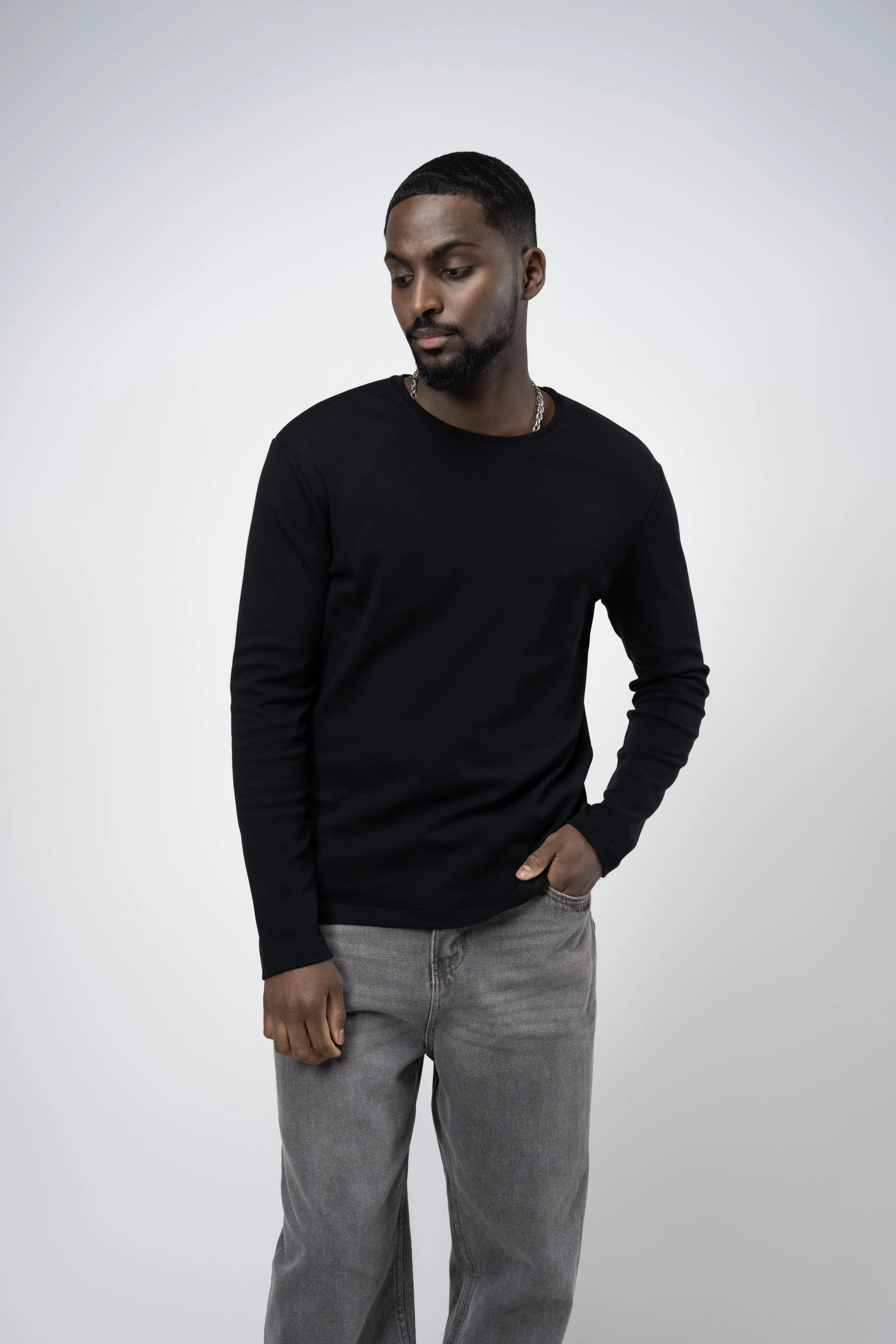 A young man with dark hair and beard dressed in a black long sleeve shirt and gray pants, standing against a plain light gray background, looking down with one hand in his pocket.