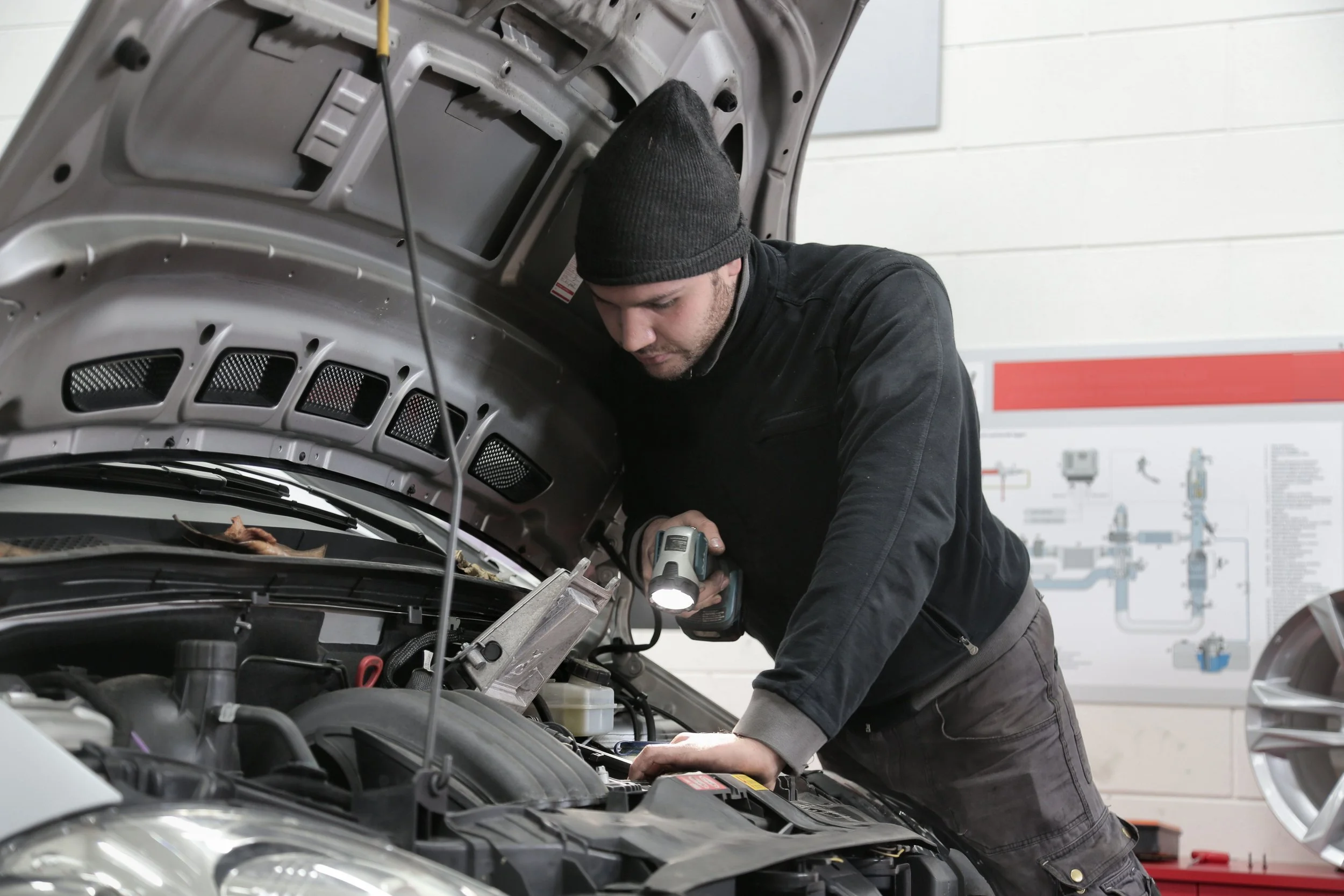 Mechanic using a flashlight to inspect a car engine beneath an open hood.