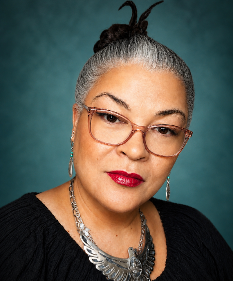 Portrait of a mature woman with gray hair styled in an updo, wearing glasses, red lipstick, earrings, and a statement necklace against a teal background.