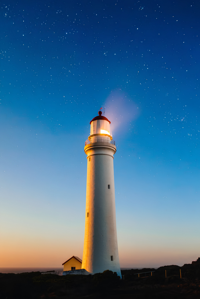 A tall white lighthouse with red trim at the top, illuminated and shining its light against a night sky filled with stars, with a small yellow building at its base and the horizon in the background.