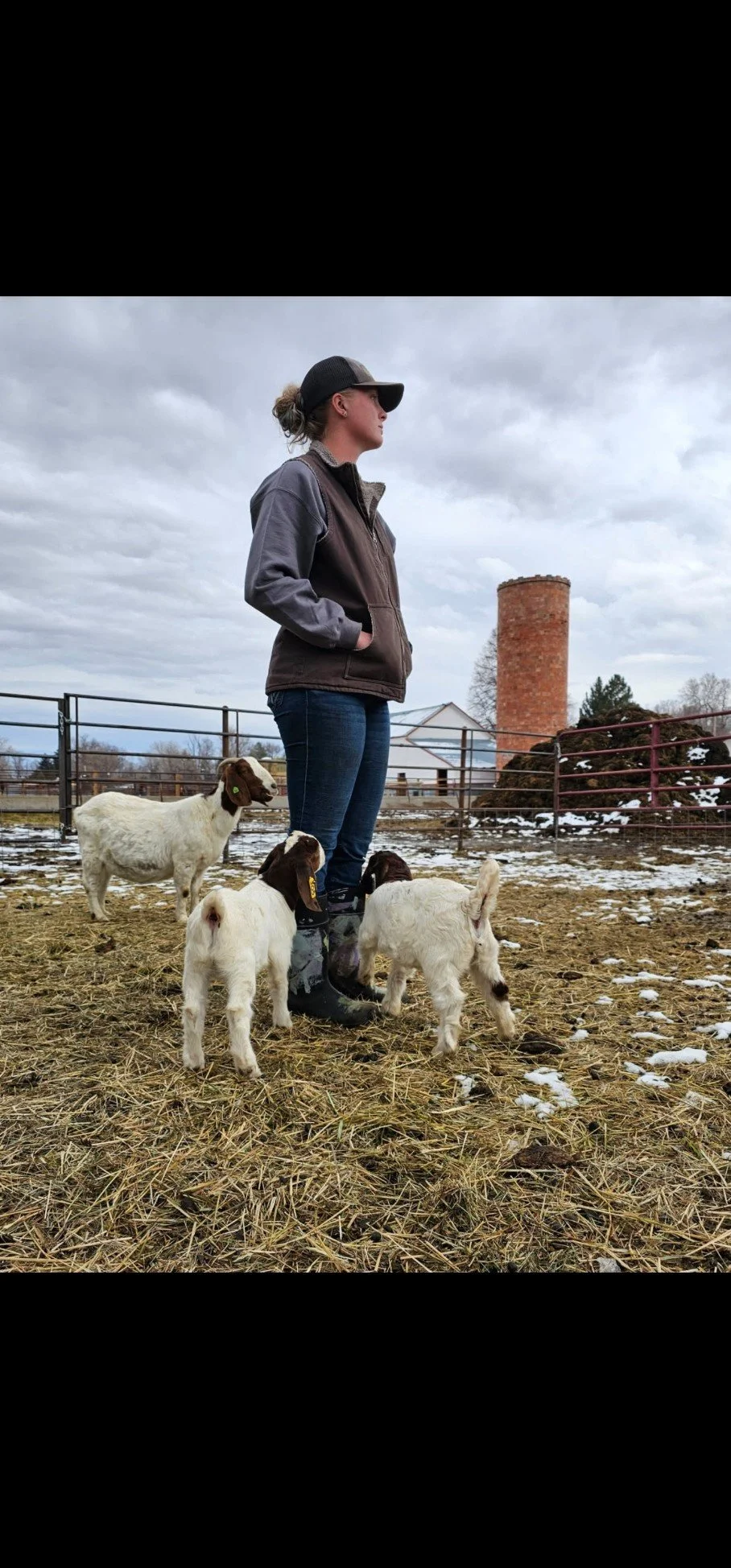 A woman standing in a muddy farmyard with three baby goats, with a barn and a silo in the background on a cloudy day.