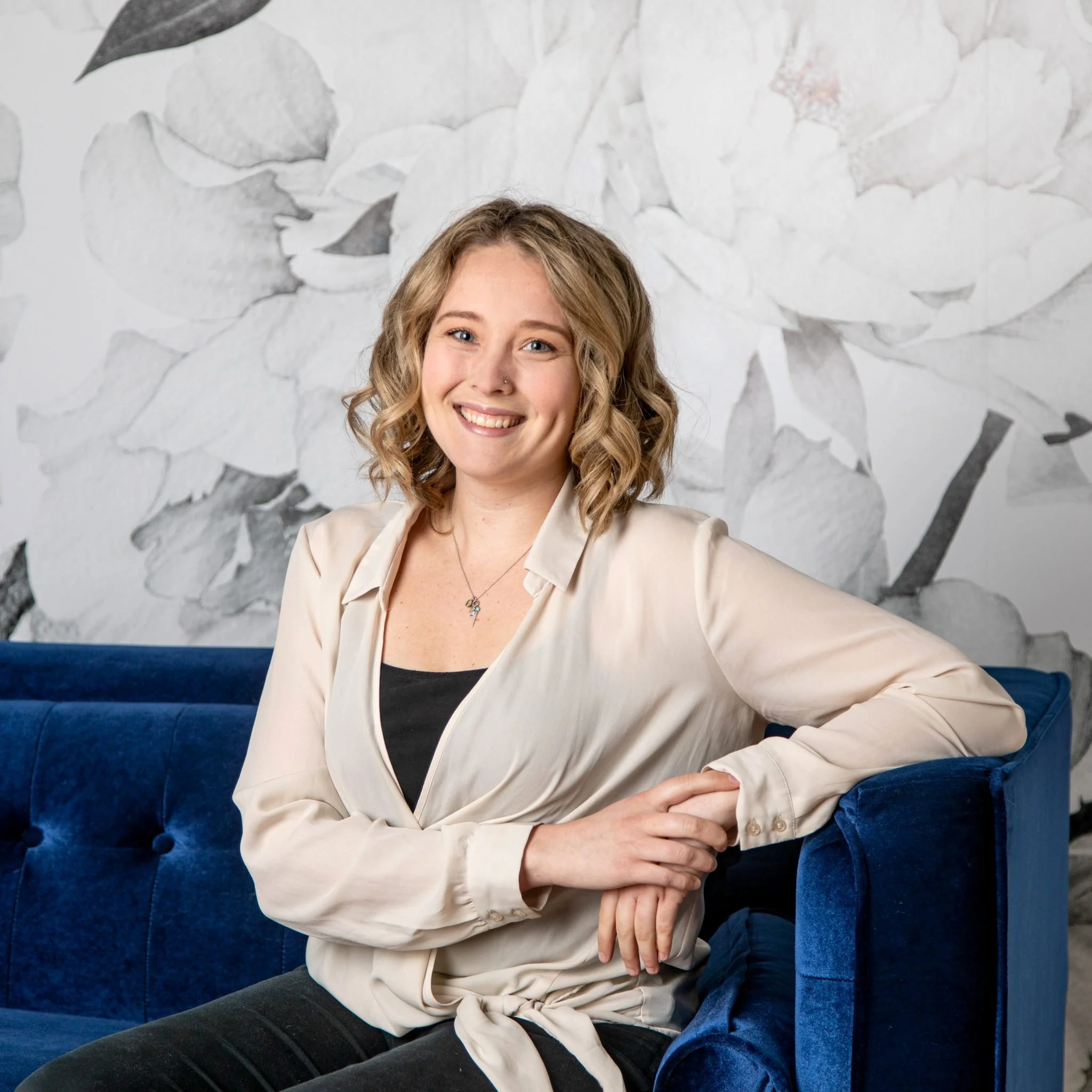 A smiling young woman with light brown curly hair and light skin wearing a beige blazer over a black top sitting on a blue velvet chair in front of a textured white wall with abstract gray and black patterns.