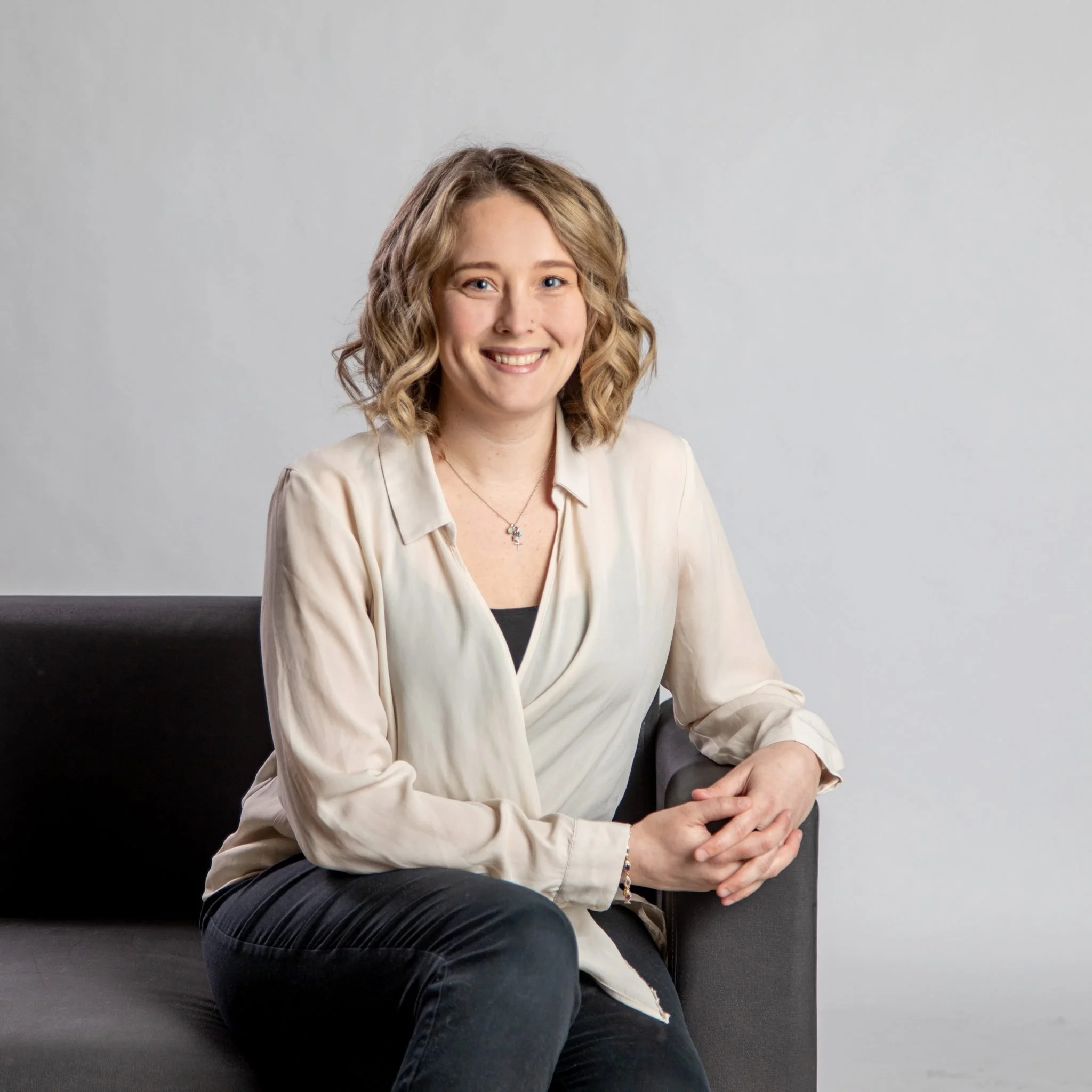 Young woman with curly blonde hair smiling in front of a plain gray background, sitting on a black couch with her arms resting on her hips.