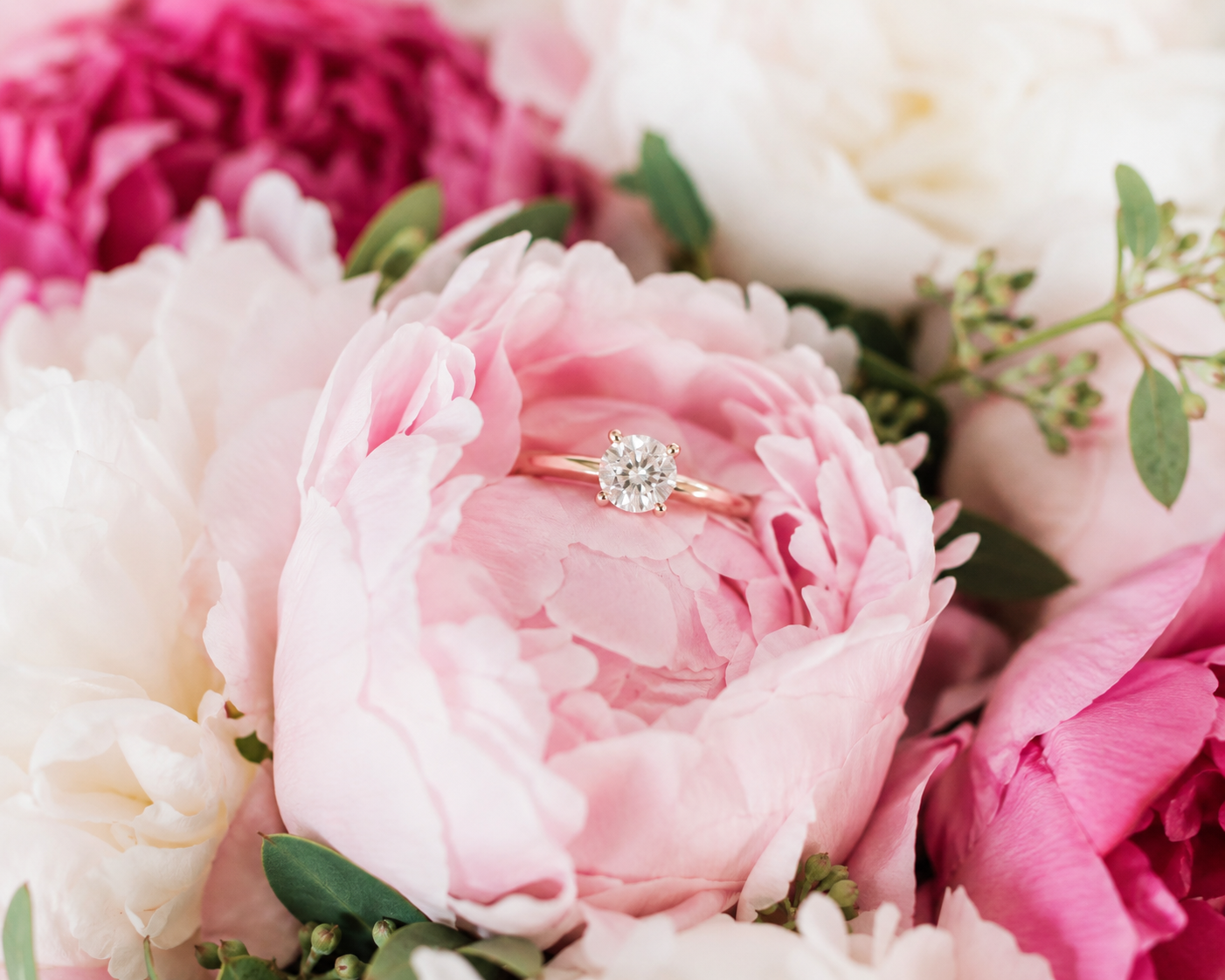 A diamond engagement ring with a round solitaire stone rests on a pink peony flower, surrounded by white and pink peonies with green leaves.