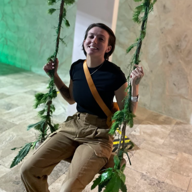 Mya, the founder of Rivers & Peonies Events, sitting on a swing made of green leaves and rope, smiling at the camera.