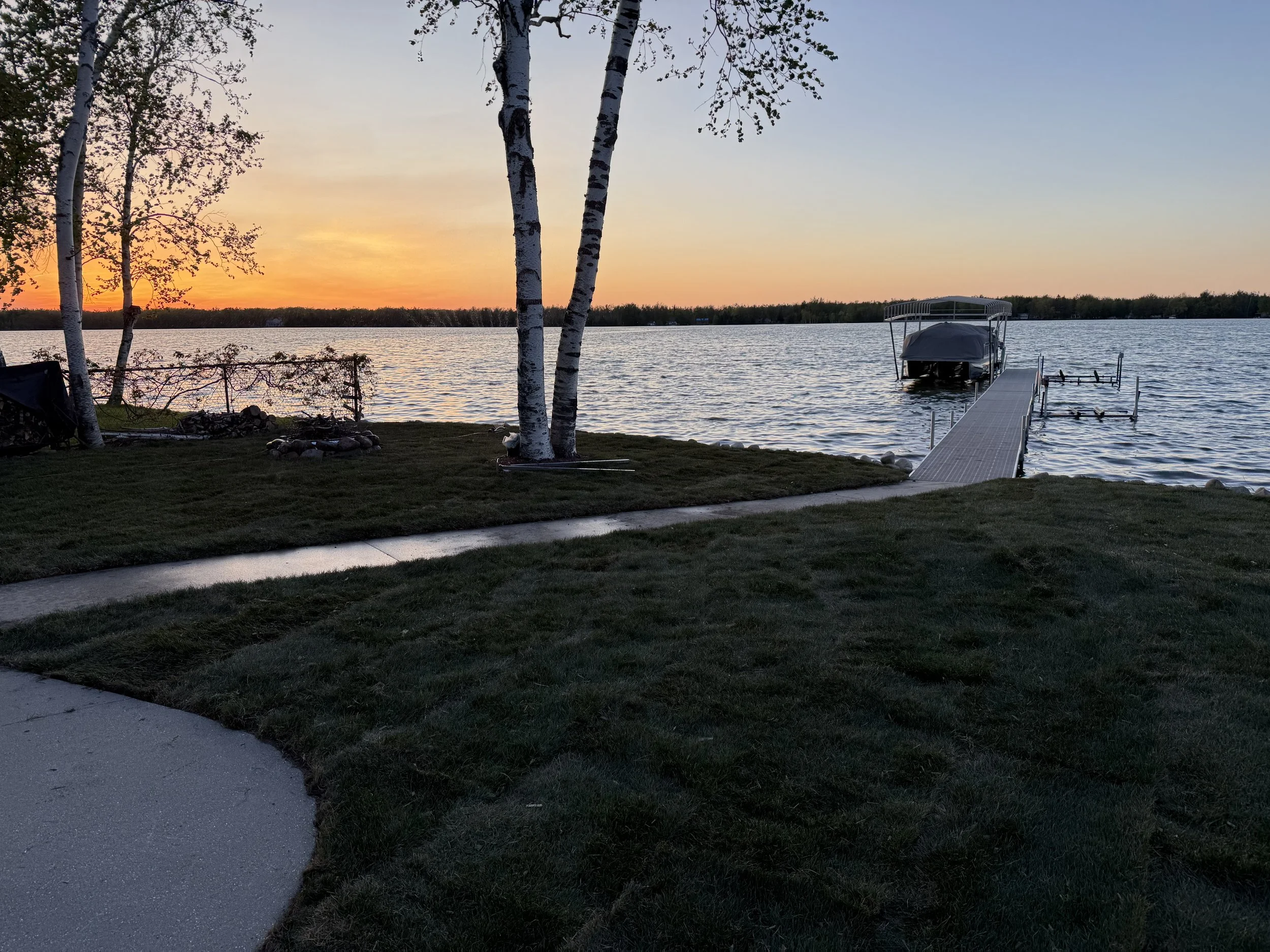A lakeside scene at sunset with a dock extending into the water, a boat covered with a tarp, trees along the shore, and a colorful sunset sky reflected on the lake surface.