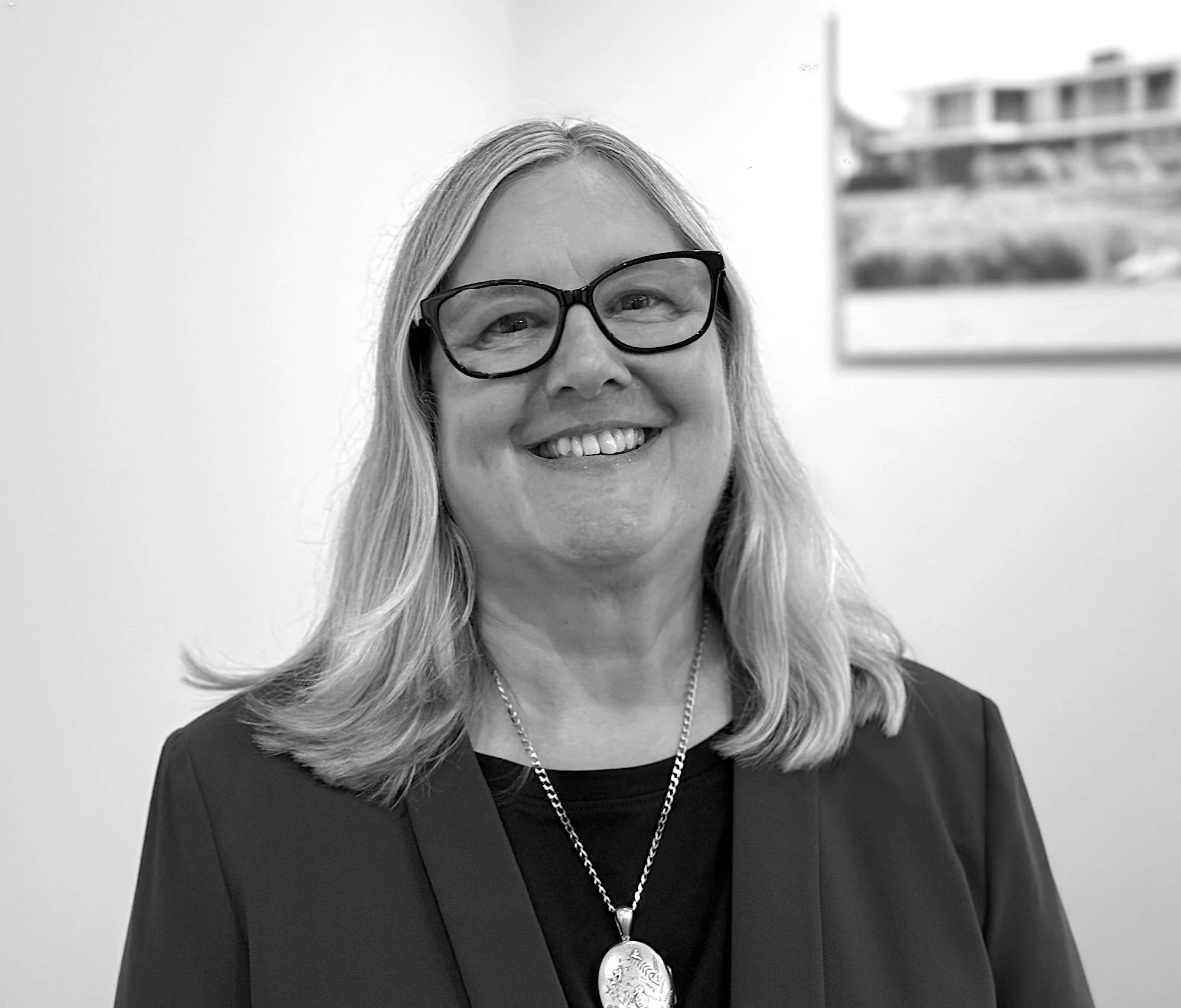 Close-up of a smiling woman with long blonde hair, wearing black glasses, a necklace with a large pendant, and a dark top, with a blurred painting or photograph on the wall behind her.