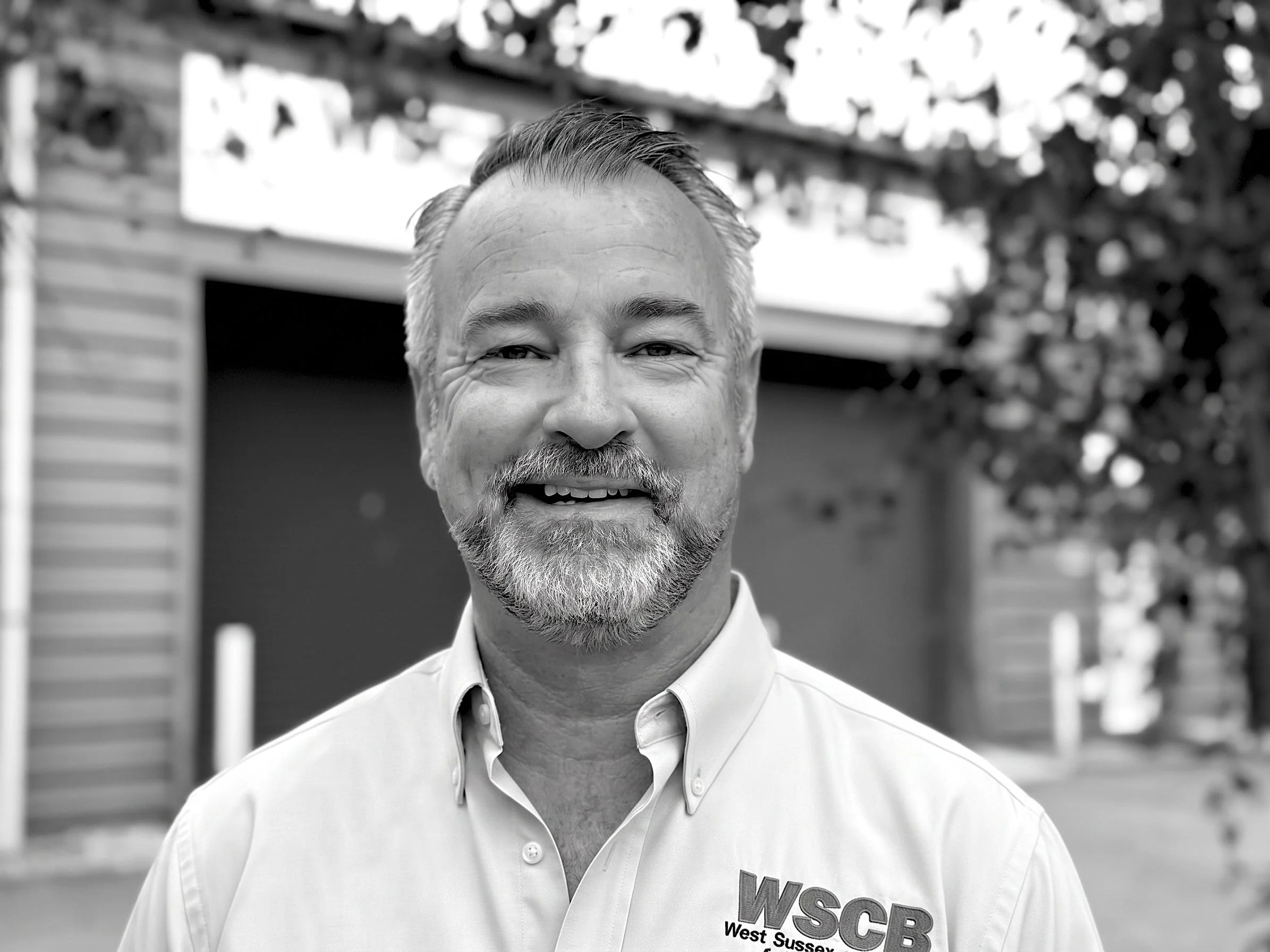 A black and white photo of a middle-aged man with a beard and short combed hair, smiling outdoors. He is wearing a white collared shirt with a logo on the chest that reads "WSCR West Sussex". The background shows a fence and trees.