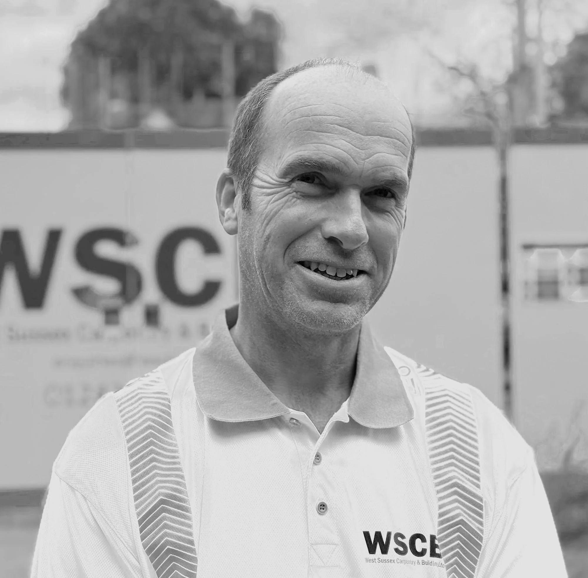 Black and white photo of a smiling man wearing a shirt with the logo 'WSCB' and text 'West Sussex Carpentry & Building Ltd.' visible on his chest, standing outdoors with blurred trees and a structure in the background.