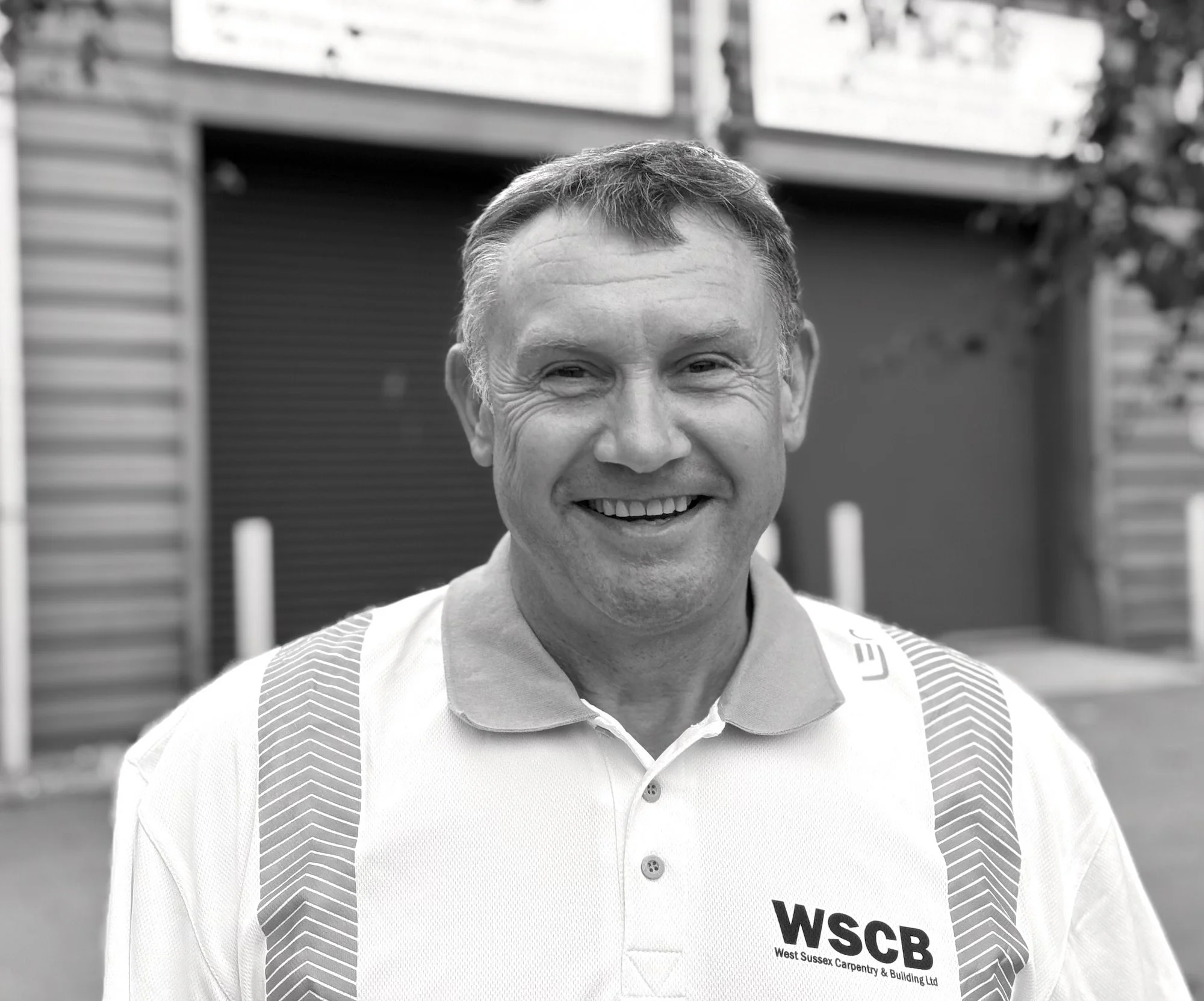 Close-up of a smiling man wearing a white collared shirt with construction straps, standing outdoors with a blurred background of houses.