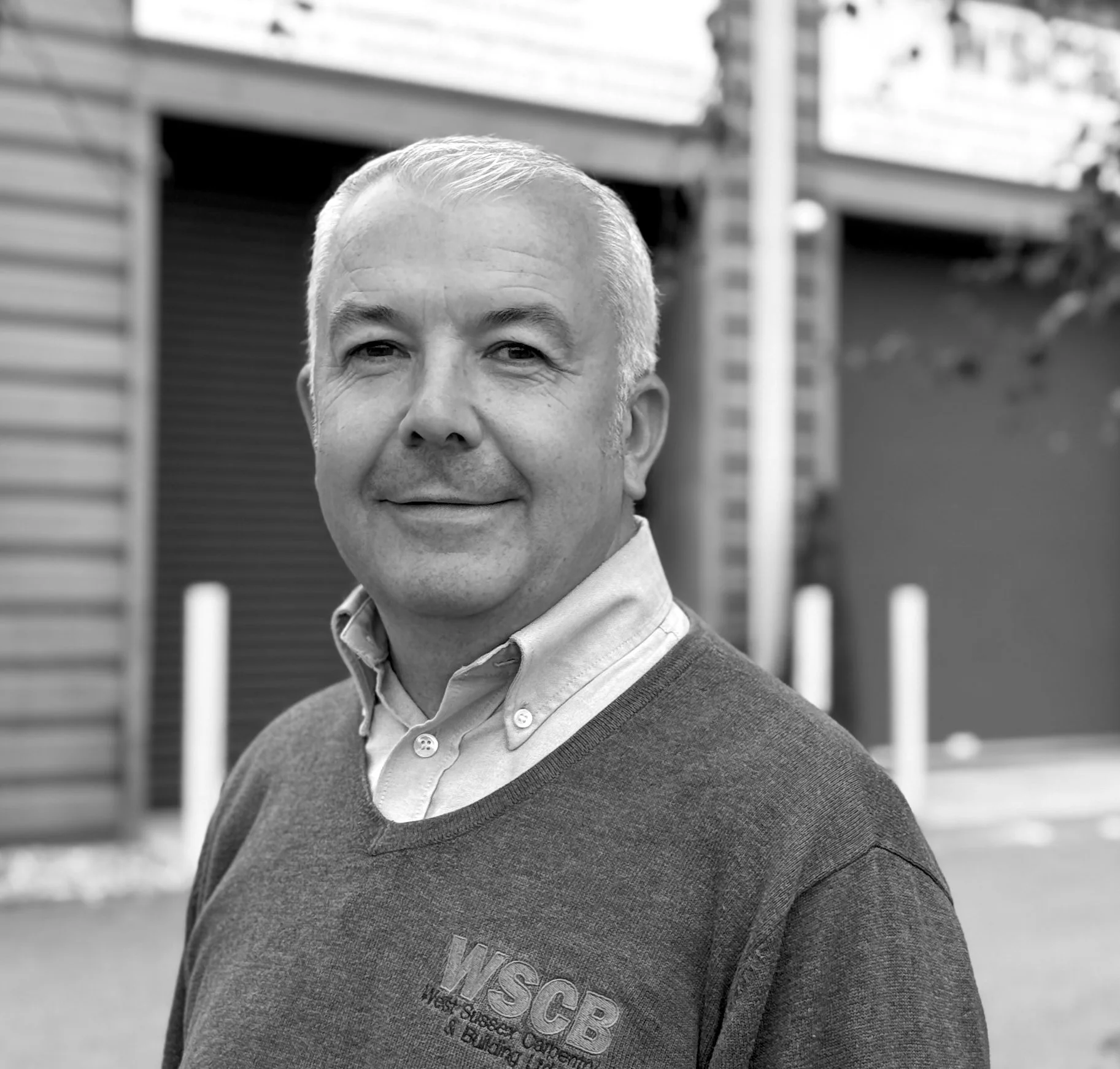 Black and white portrait of a man with short gray hair, wearing a collared shirt and a sweater with the letters 'WSCB' on it, standing outdoors in front of a house with horizontal siding and a large chimney.