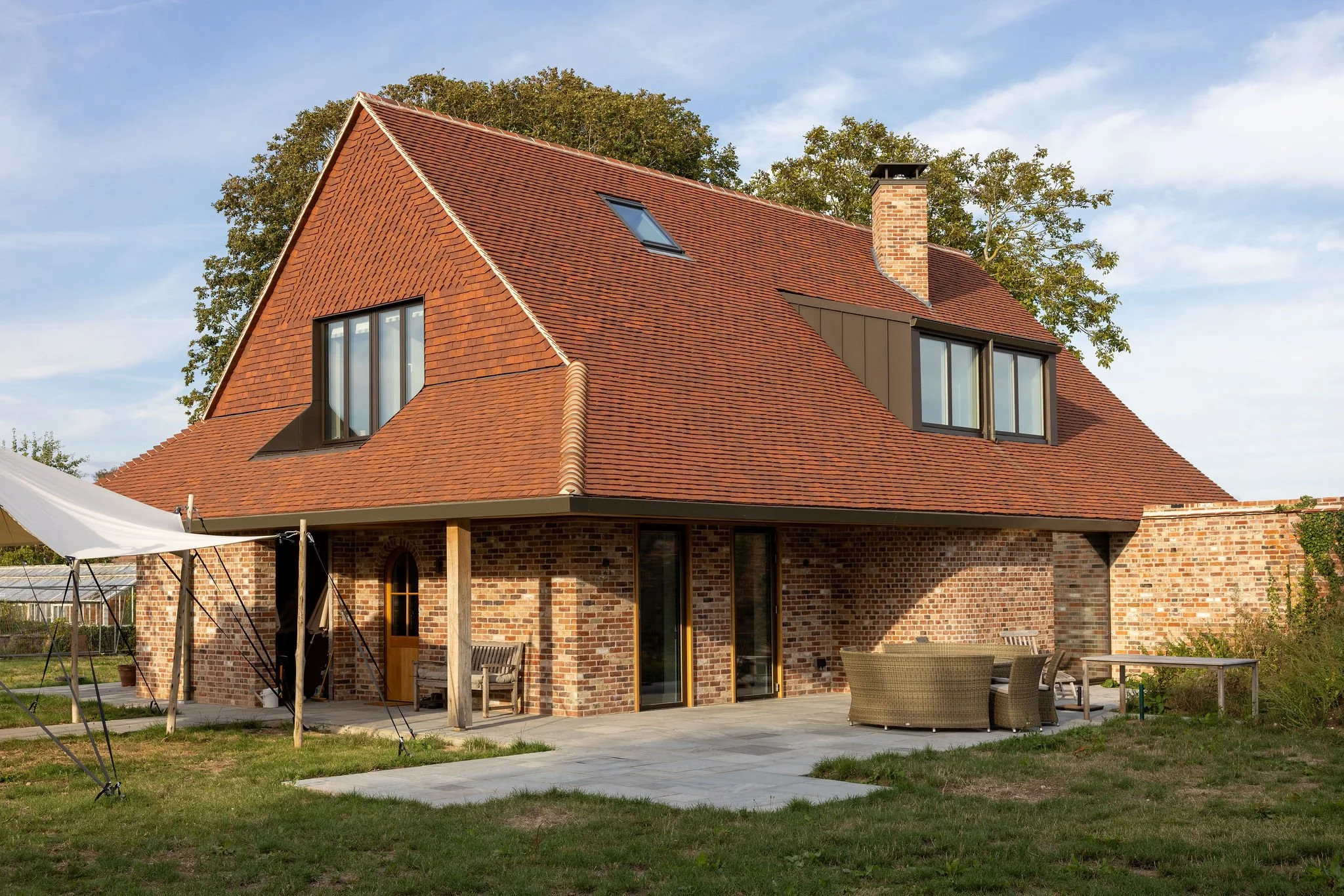 A brick house with a steep red tile roof, two large windows, and a skylight, surrounded by trees with green and brown leaves, and a gravel yard with fallen leaves.
