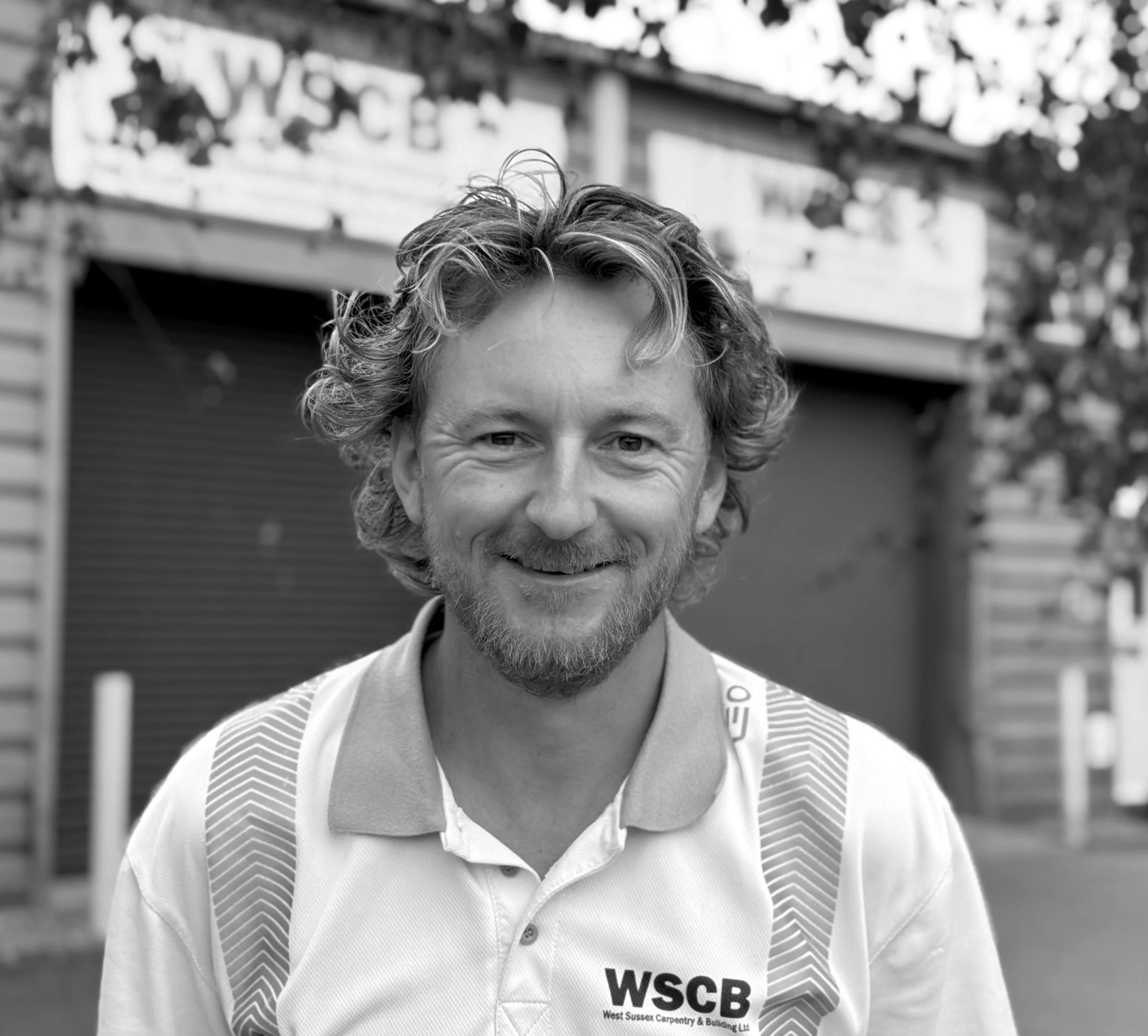 Black and white photo of a smiling man with curly hair and a beard, wearing a polo shirt and a vest with a company logo, outdoors with a blurred fence and trees in the background.