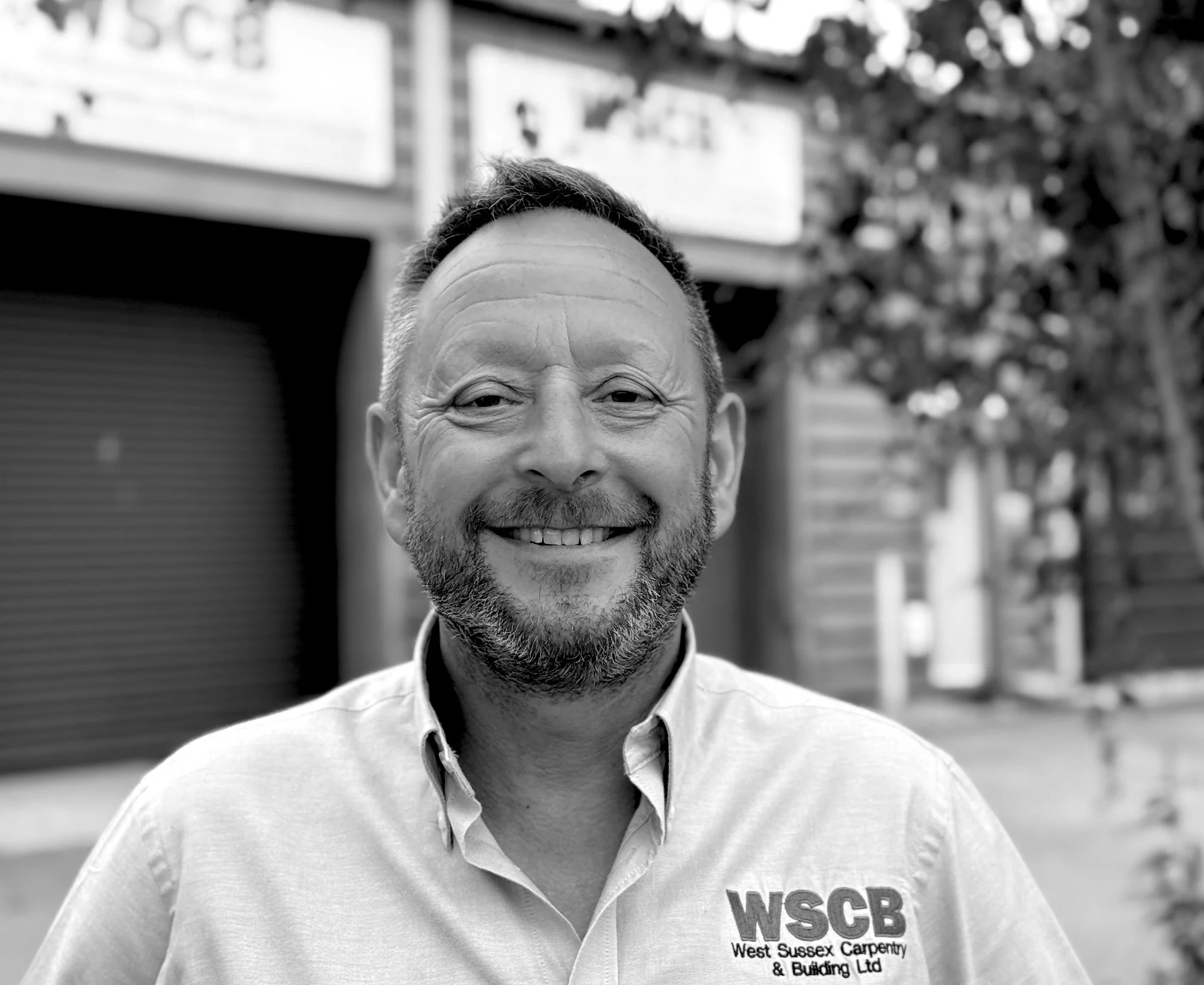 Black and white photo of a smiling man with short, styled hair and a beard, wearing a collared shirt with a logo that says 'WSCB West Sussex Carpentry & Building Ltd.' The background is blurred with trees and a building.