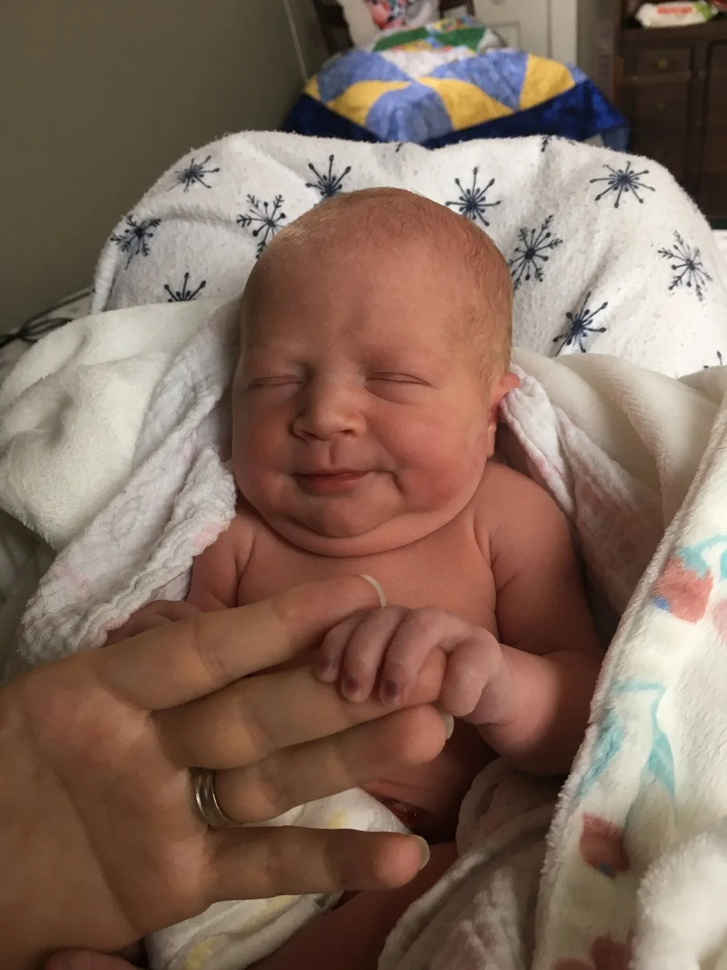 A newborn baby with a happy, smiling expression, lying on a bed wrapped in white blankets. An adult's hand gently touches the baby's hand. The background shows a bed with a warm, colorful quilt.