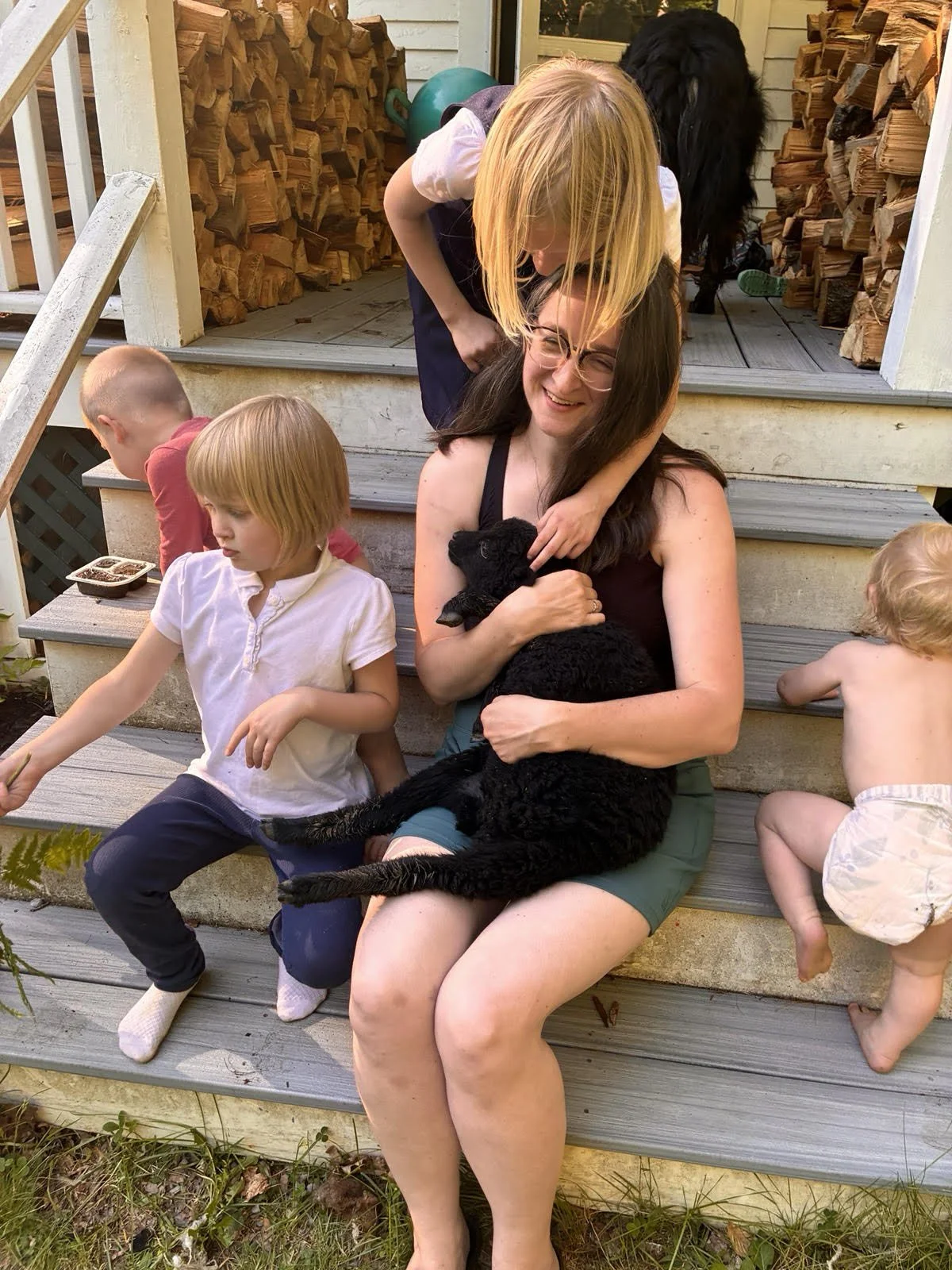 A woman sitting on outdoor steps holding a black lamb, smiling warmly as a girl leans over her head. Several children around her, one with a bowl of food, and multiple stacked firewood logs in the background.