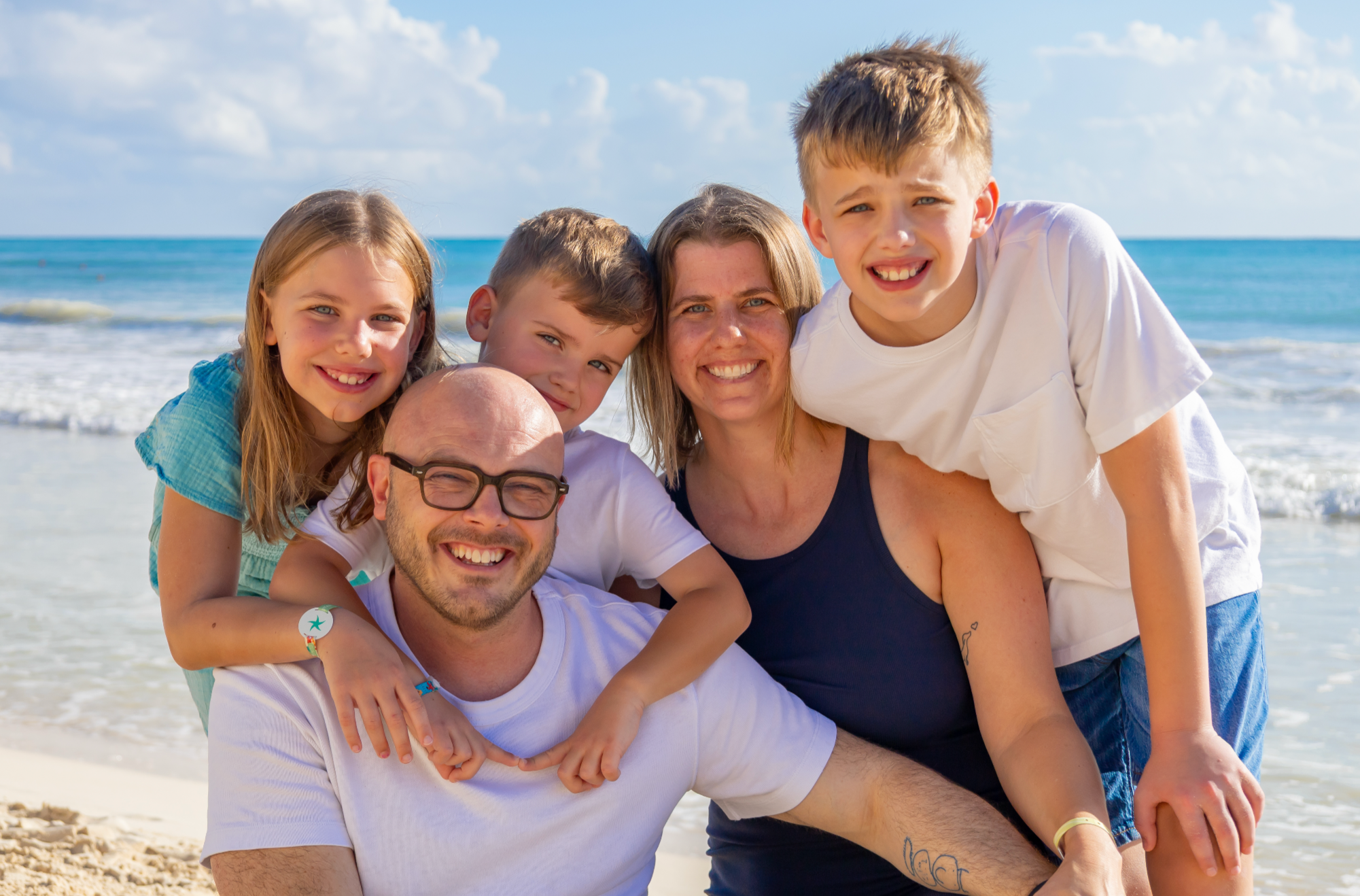 A family of five enjoying a day at the beach, smiling, standing close together with the ocean and a cloudy sky in the background.