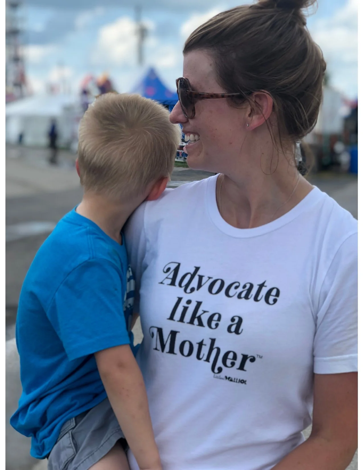A woman with brown hair in a bun, wearing sunglasses and a white T-shirt that reads "Advocate like a Mother," is smiling and holding a young boy with blonde hair in a blue T-shirt. They are outdoors at a fair or festival with tents and people blurred in the background.