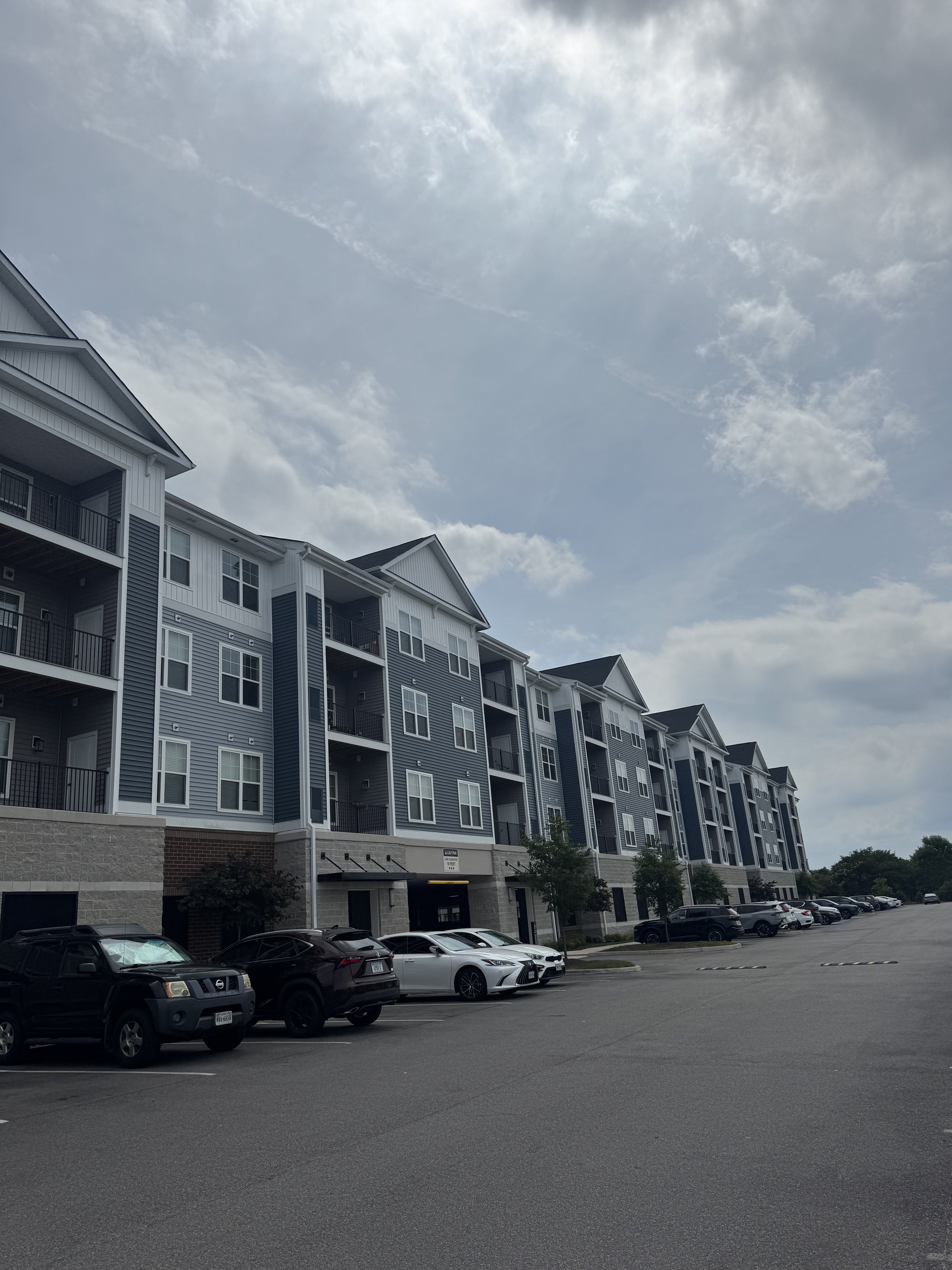 Multi-story apartment building with balconies, parked cars in a parking lot, and a partly cloudy sky above.
