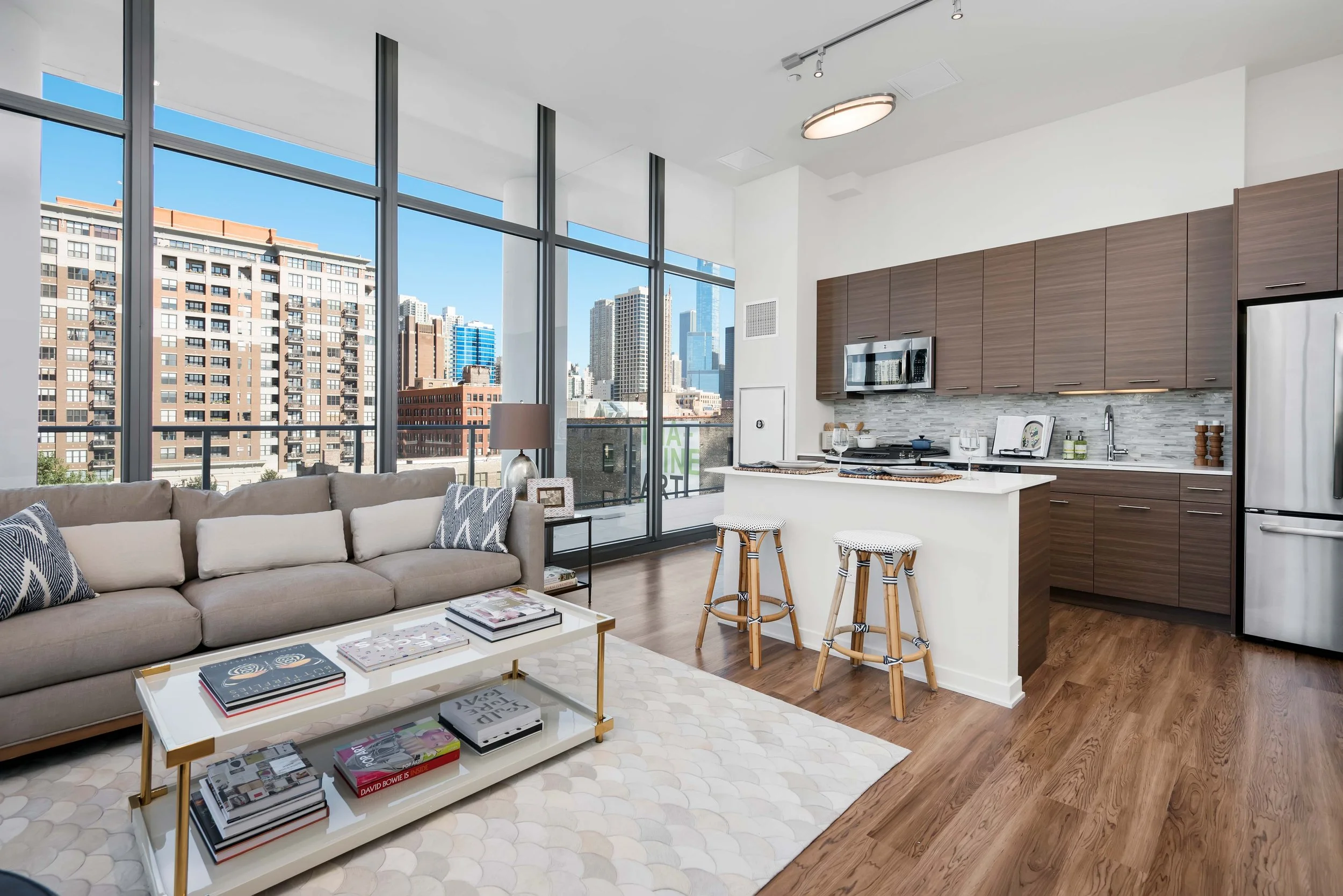 Living room with large windows, city skyline view, beige sofa with patterned pillows, white coffee table with magazines, kitchen with brown cabinets, stainless steel appliances, and bar stools at a white counter.