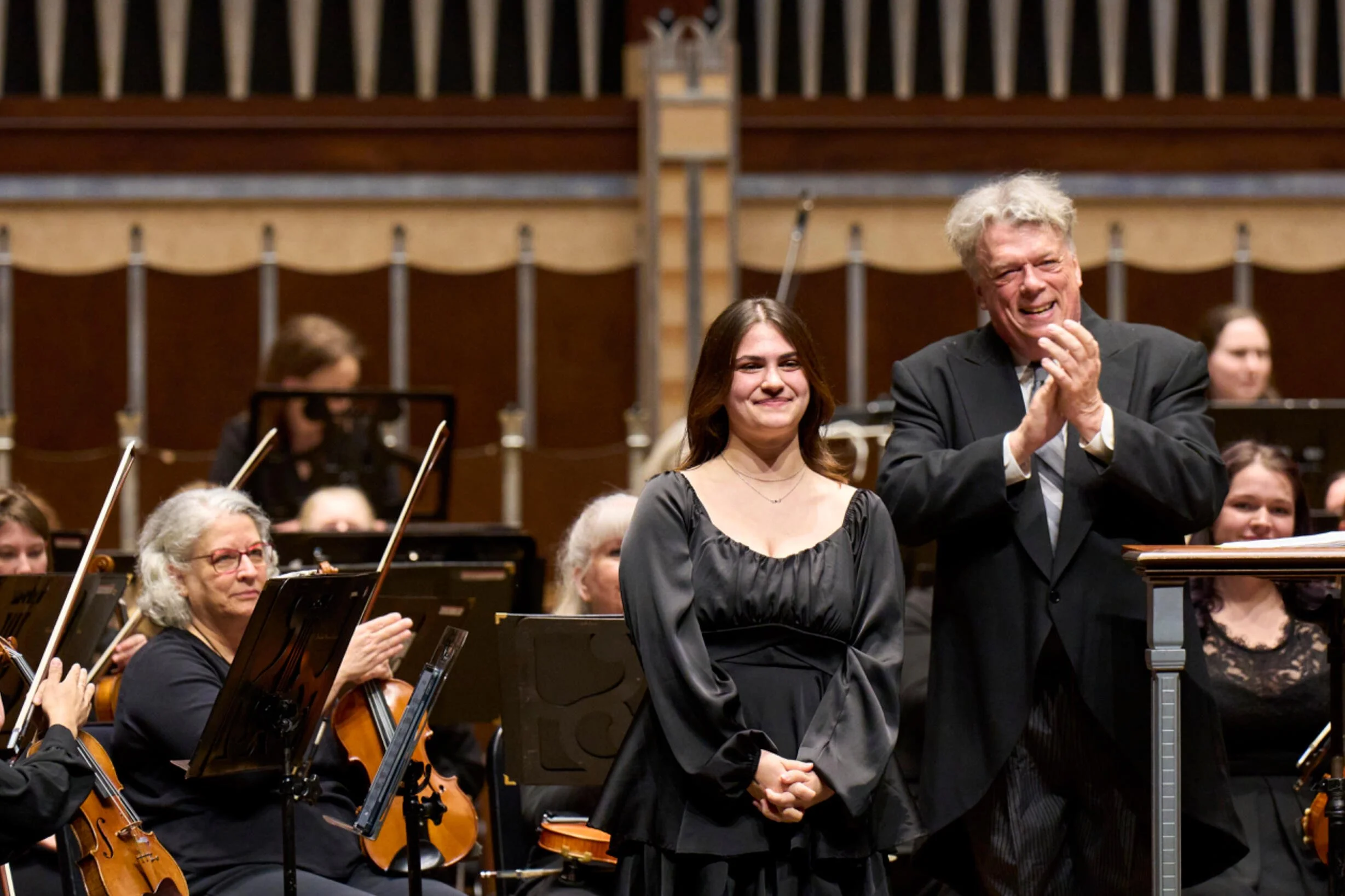 An orchestra conductor and a young woman are smiling on stage after a performance, with orchestra members playing violins and cellos around them in a concert hall.