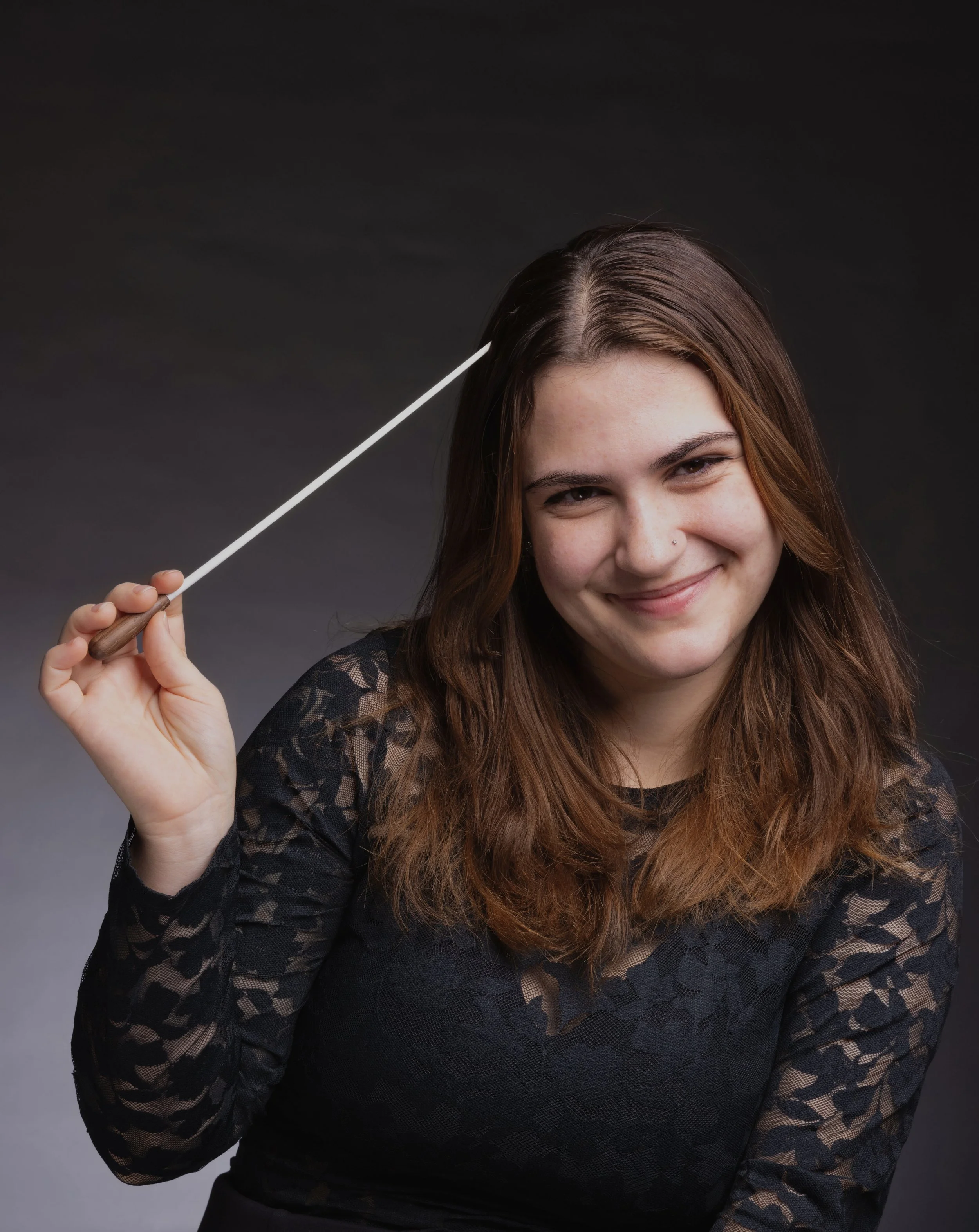 A young woman with long brown hair and a nose piercing smiling and holding a pointer stick, dressed in a black lace top, standing against a dark gray background.