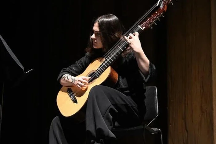 A woman with long dark hair playing an acoustic guitar on a stage, seated on a chair with a dark background.
