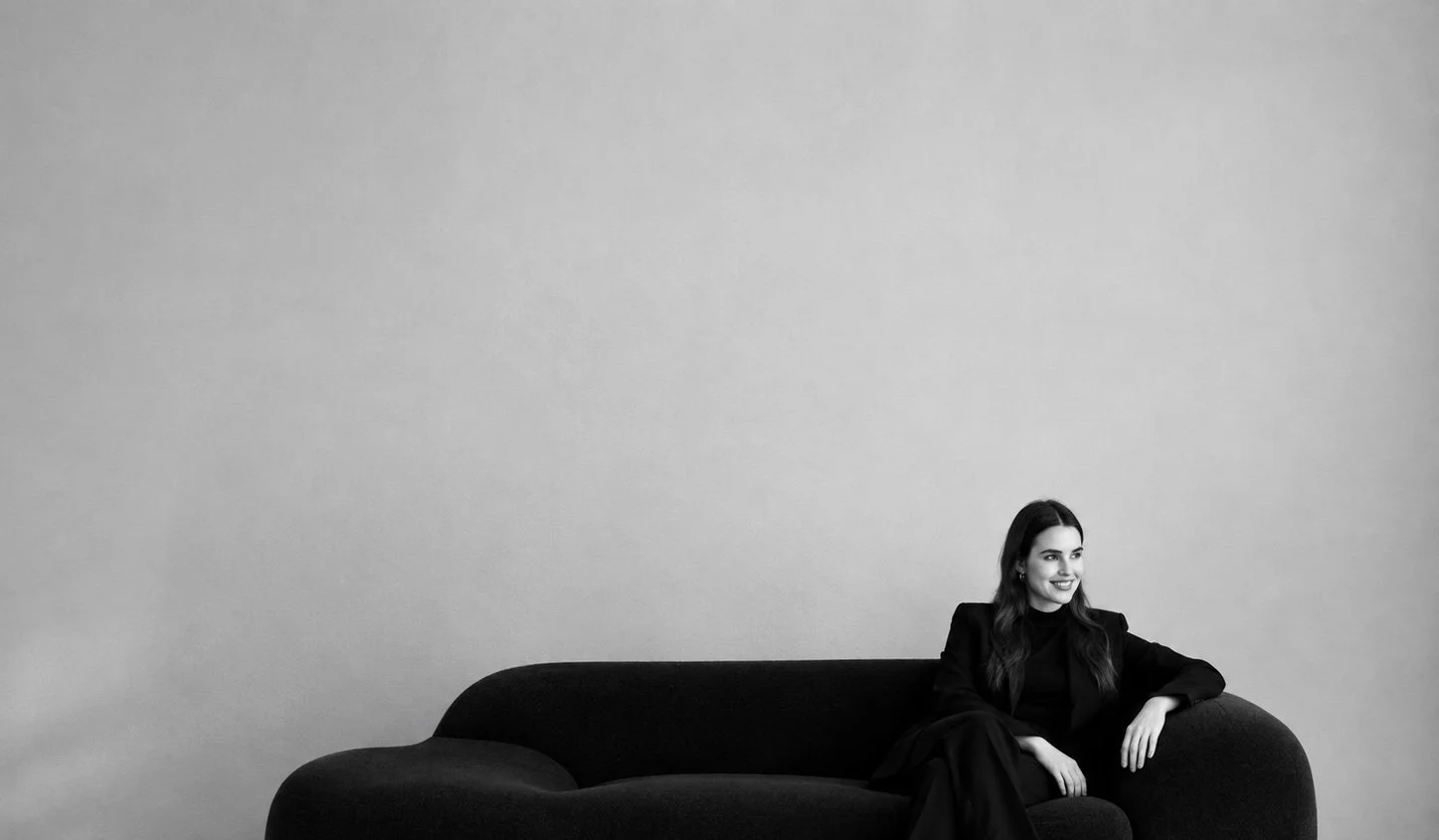Schwarz-weiß Foto einer lachenden Frau in dunkler Kleidung, die auf einer modernen schwarzen Couch sitzt, vor einer leeren hellgrauen Wand.