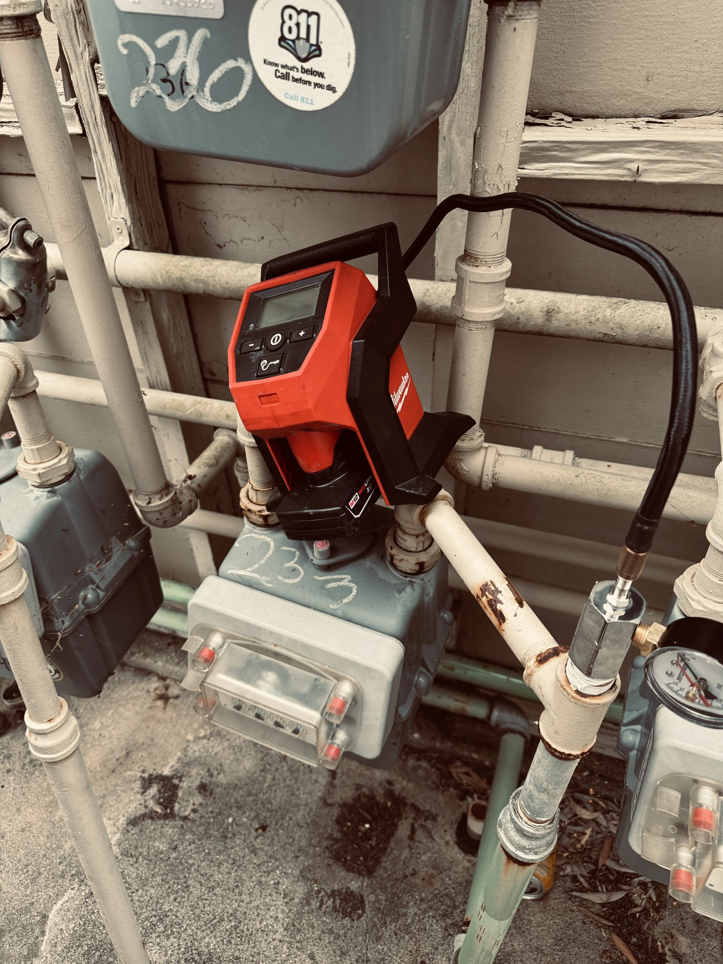An outdoor utility area with pipes and meters, including a red Milwaukee thermal imaging device resting on a pipe, and various electrical and plumbing components.