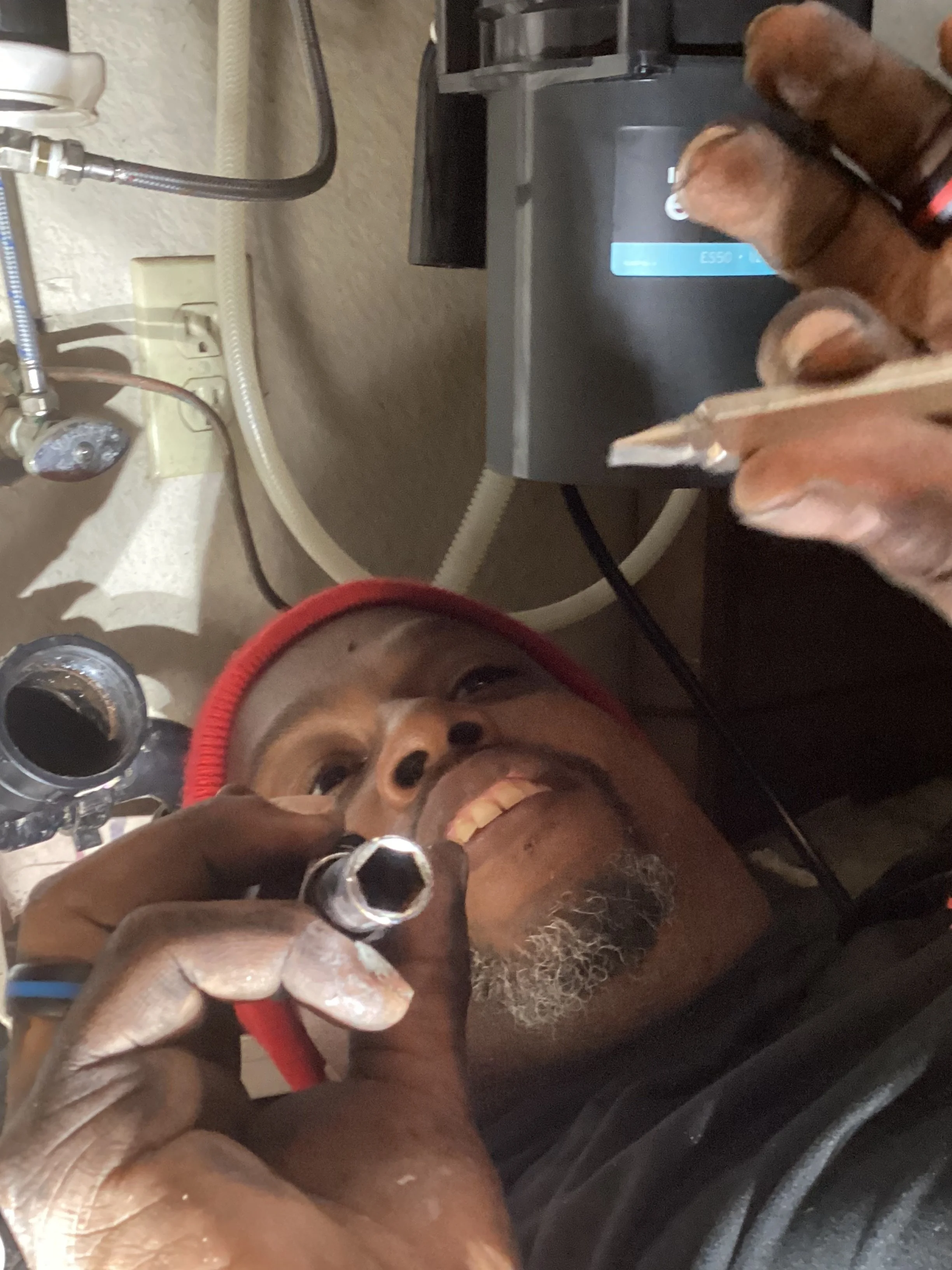 A man lying on a table or bed, smiling, with a red headband, holding a metal tool, with a dog gently touching his face with its paw, in a setting with medical or grooming equipment, including a sink, hoses, and a machine.