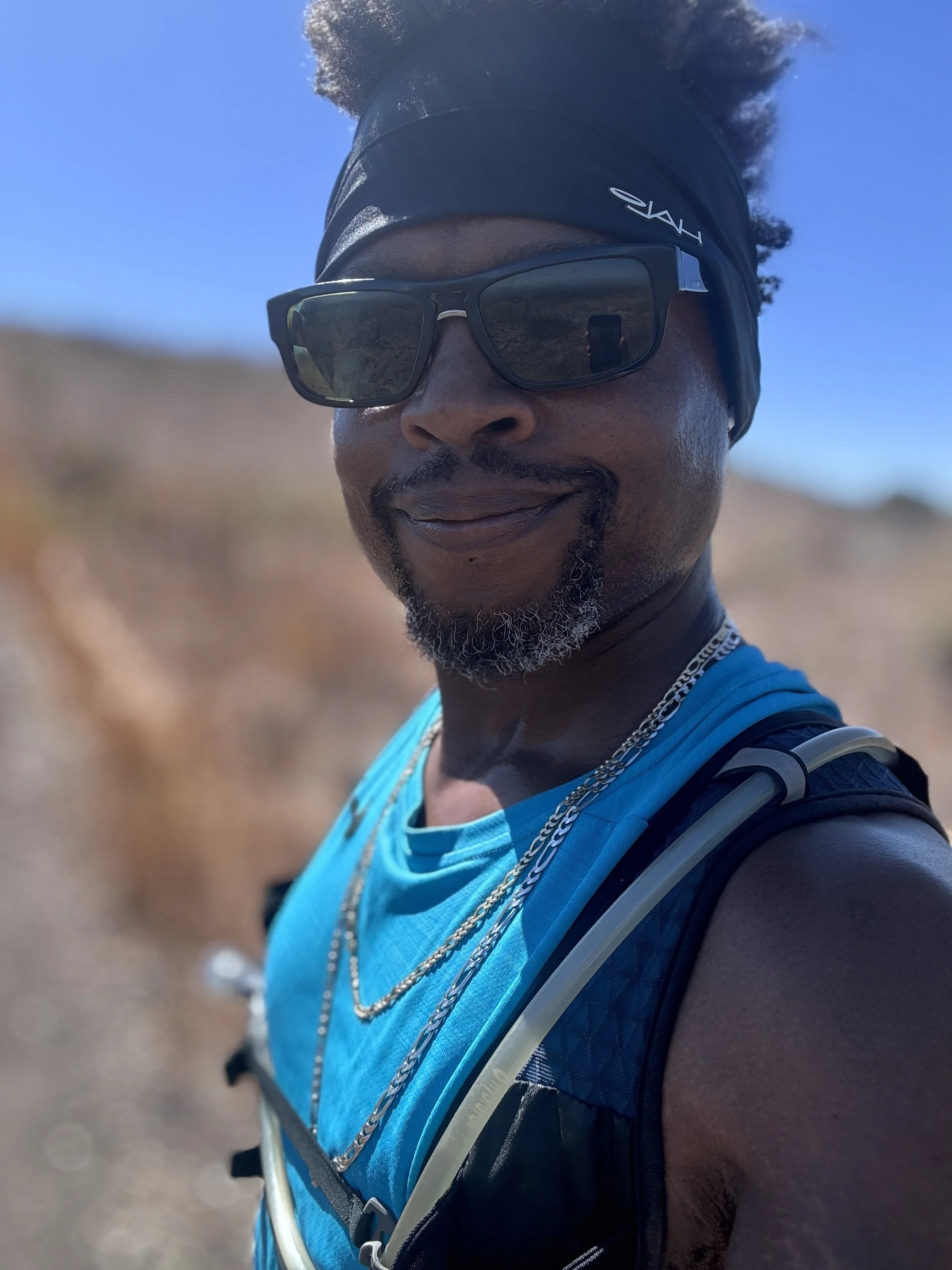 Close-up of a man wearing Prada sunglasses, a black headband, and a blue nike sports shirt, smiling outdoors on a sunny day.