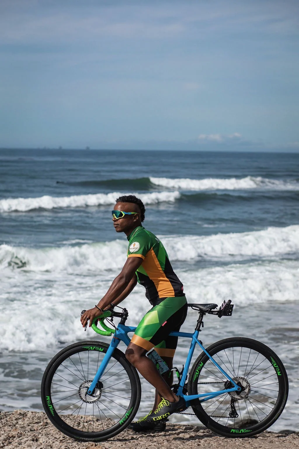 A man wearing sunglasses and a green, black, and orange cycling outfit riding a blue bicycle along the beach with ocean waves in the background.