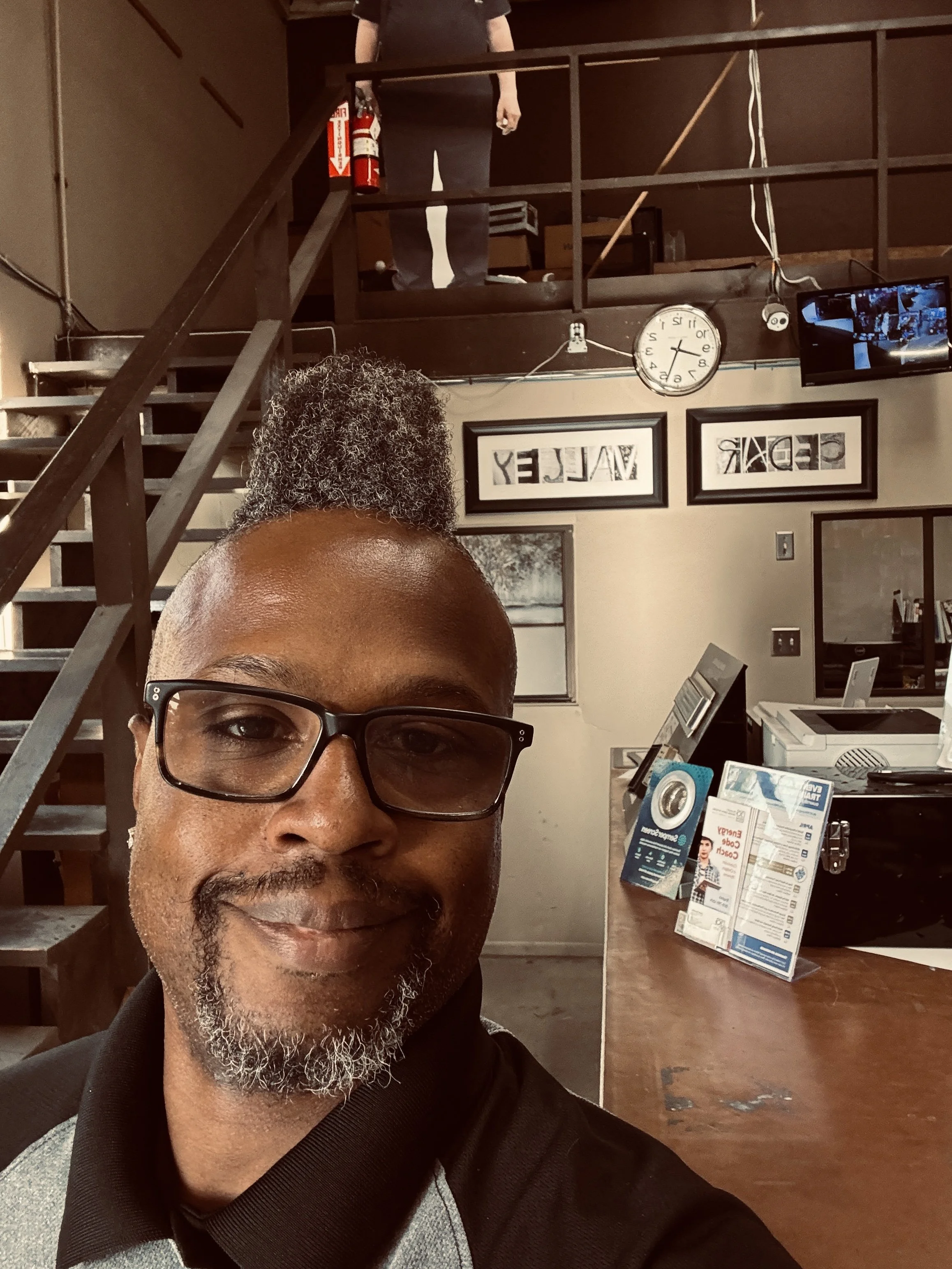 A man with glasses and a beard taking a selfie in a coffee shop or office with wooden stairs, framed pictures, a wall clock, and a TV in the background.